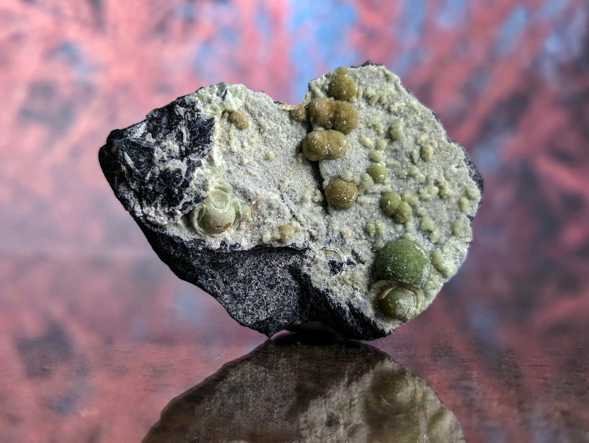 A heart-shaped rock specimen featuring a dark, rough-textured base with clusters of small, rounded greenish-yellow mineral formations.