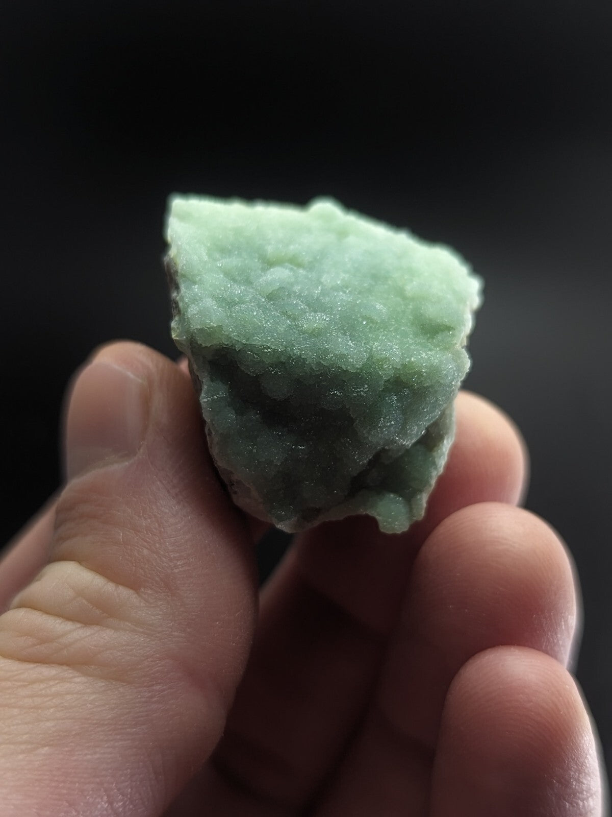 A hand holds a rough, pale green mineral specimen with a textured, crystalline surface.