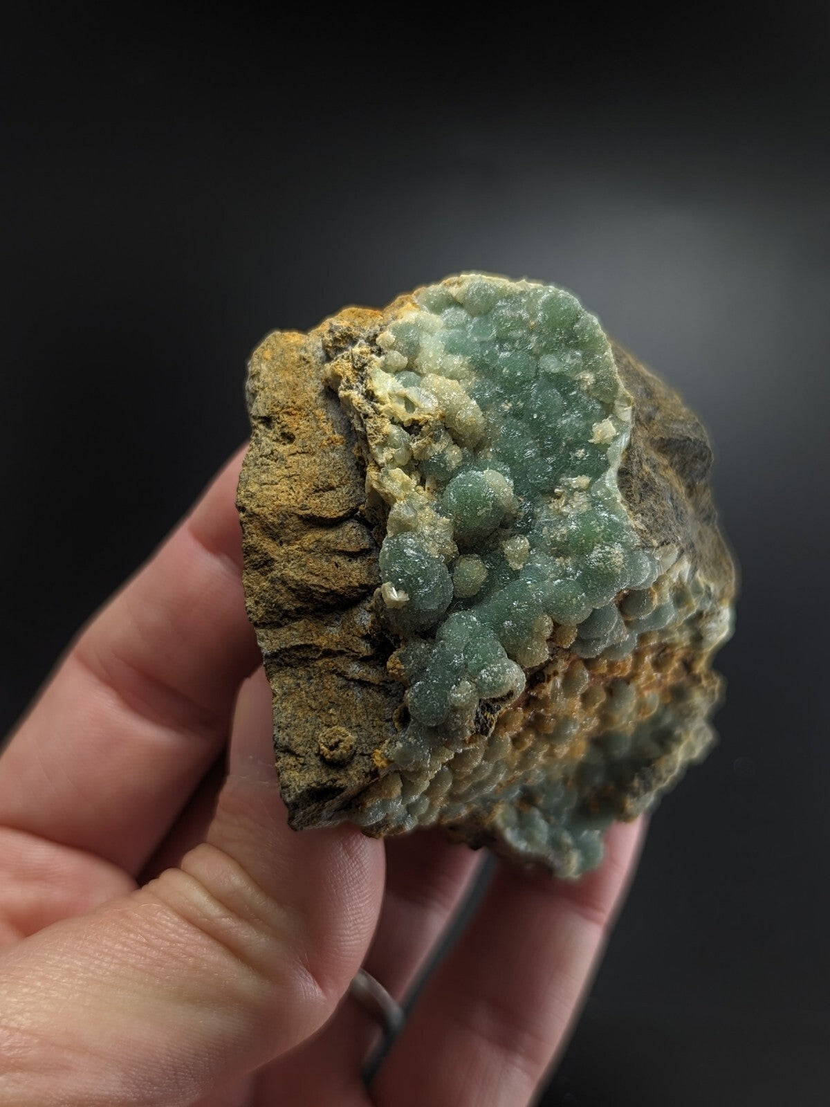 A hand holds a rough, earthy-textured mineral specimen featuring clusters of translucent green crystals growing from a brownish-yellow matrix.