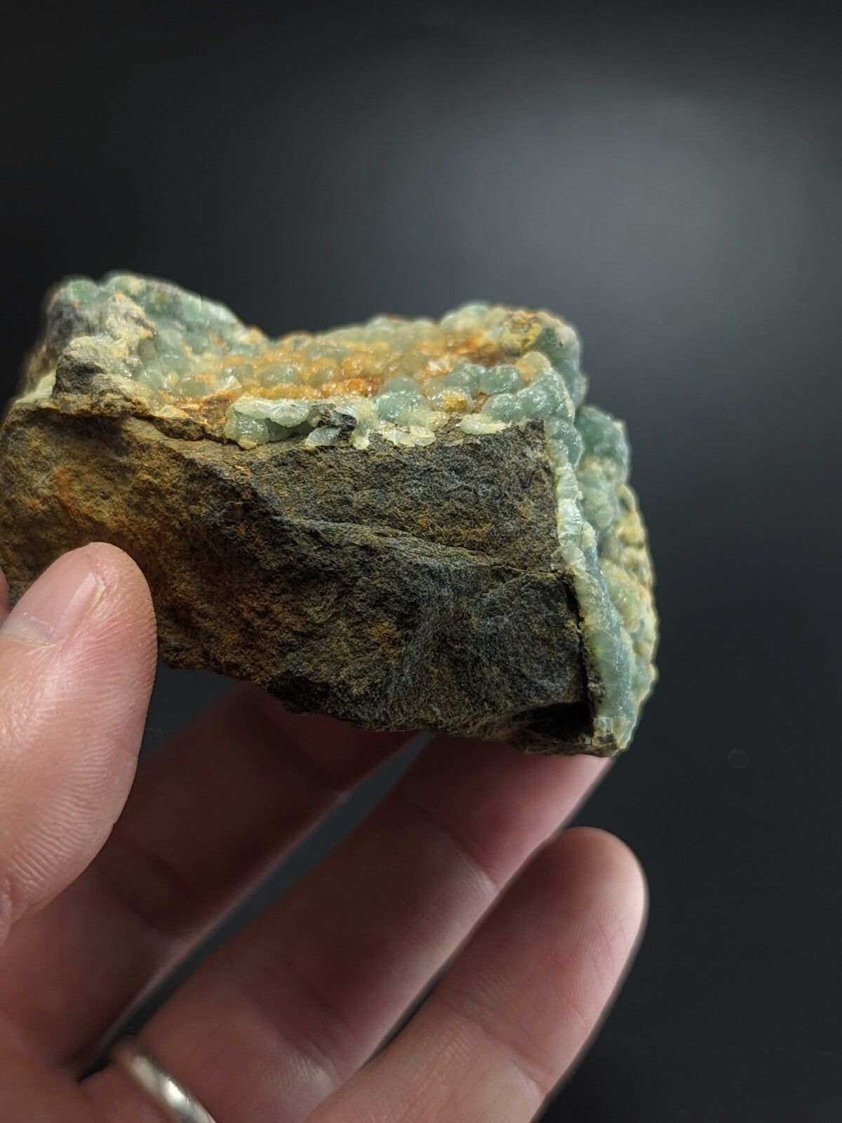 A hand holds a rough, irregularly shaped piece of mineral with translucent light green crystals embedded in a darker brown and gray matrix.