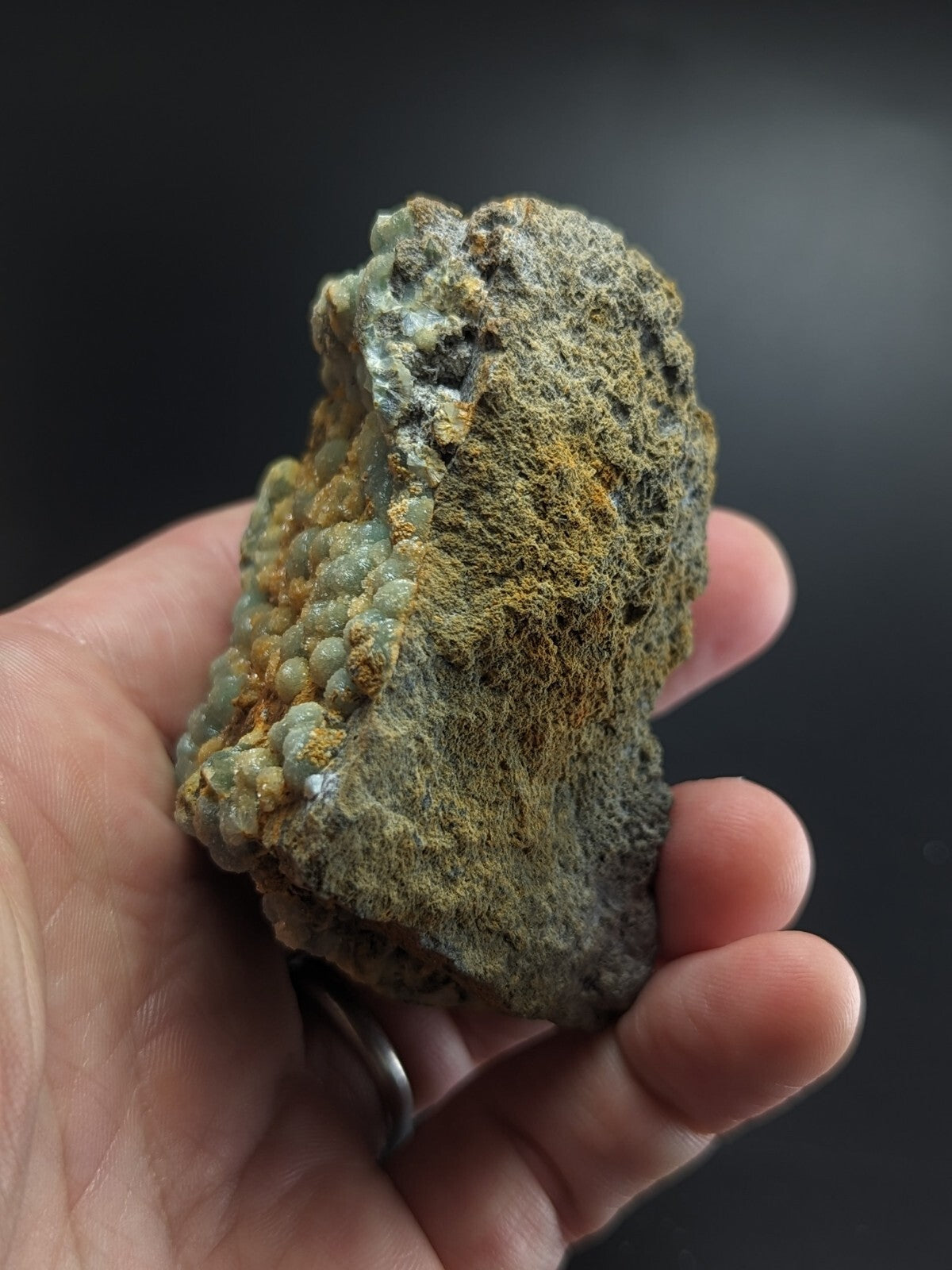 A hand holds a rough, irregularly shaped mineral specimen featuring clusters of translucent greenish-blue nodules and patches of earthy yellow-orange crust.