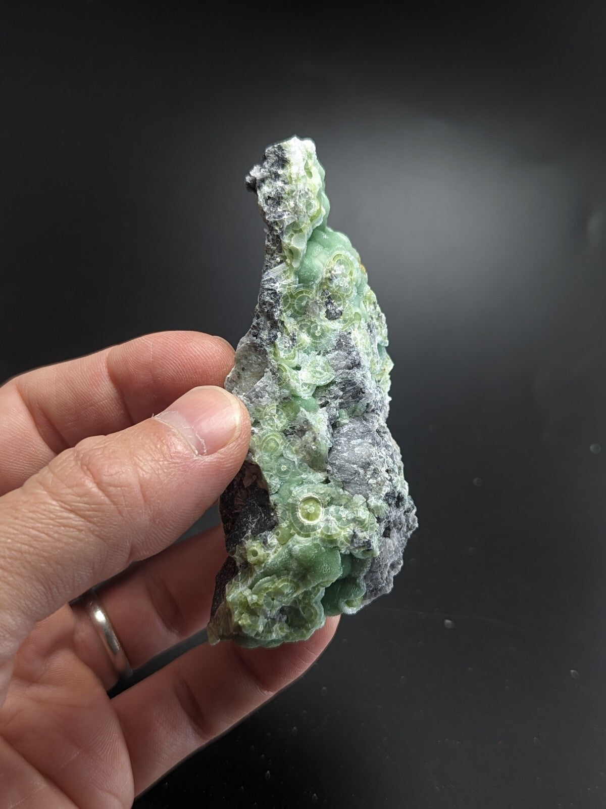A hand holds a rough, irregularly shaped green and gray mineral specimen with translucent, bubble-like formations.