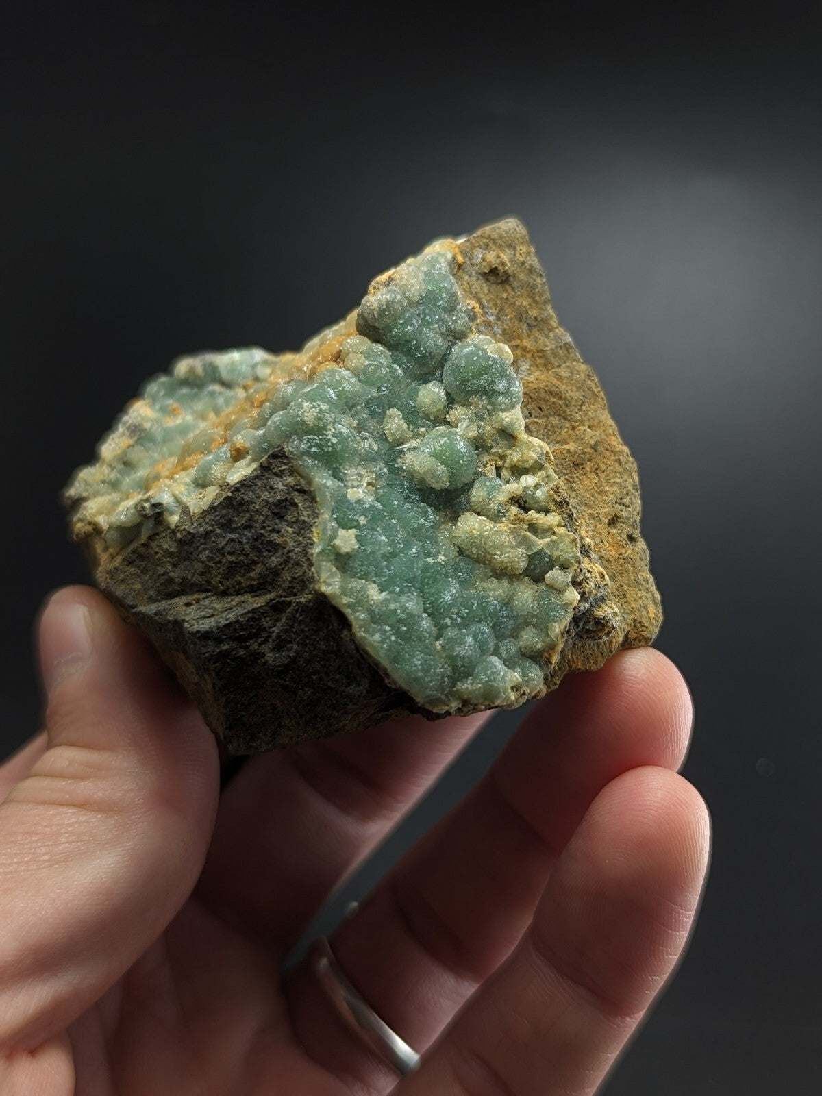 A hand holds a rough, earthy brown rock specimen featuring clusters of translucent light green crystals with subtle blue undertones.