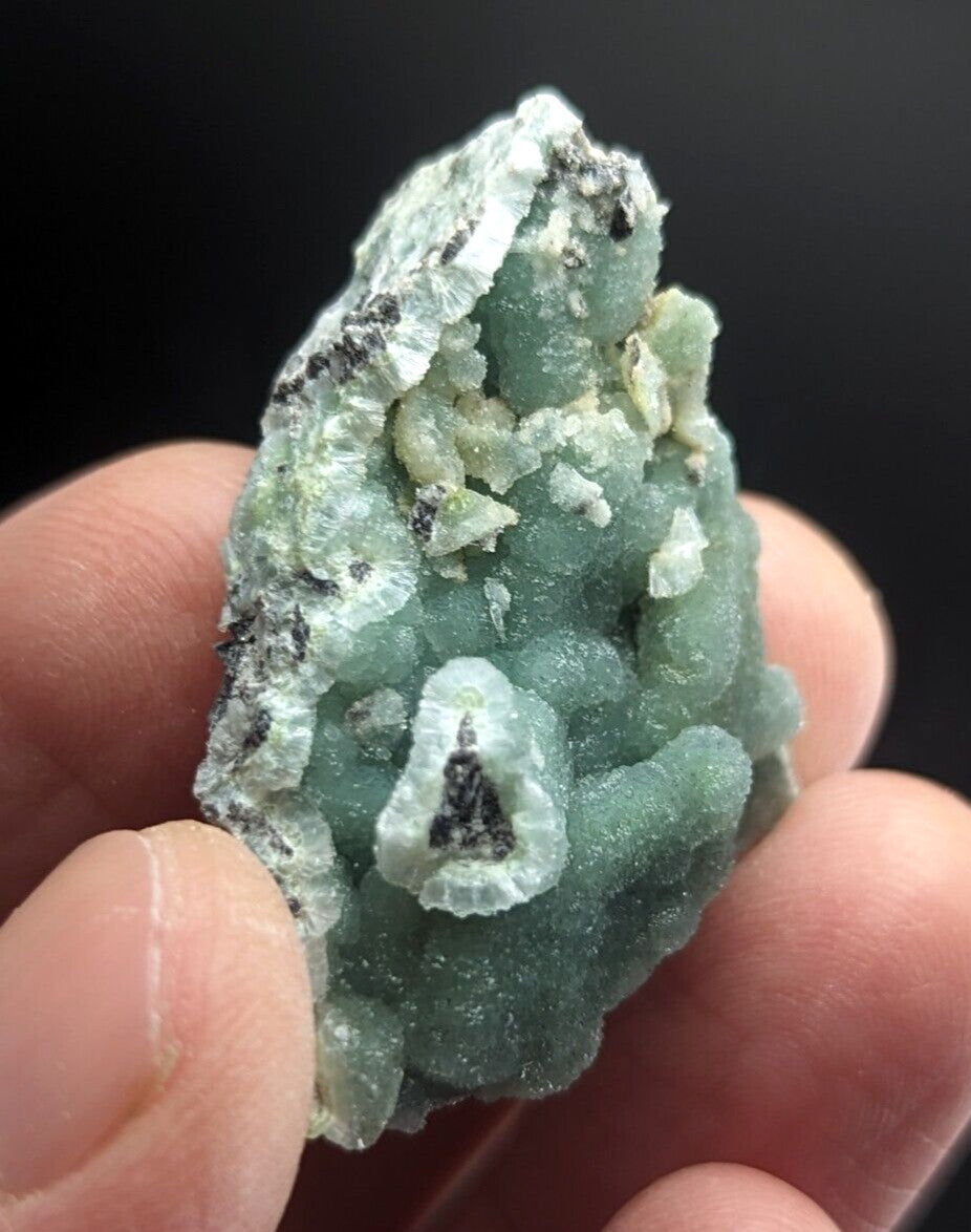 A hand holds a translucent green mineral specimen with intricate crystalline formations and dark speckles.