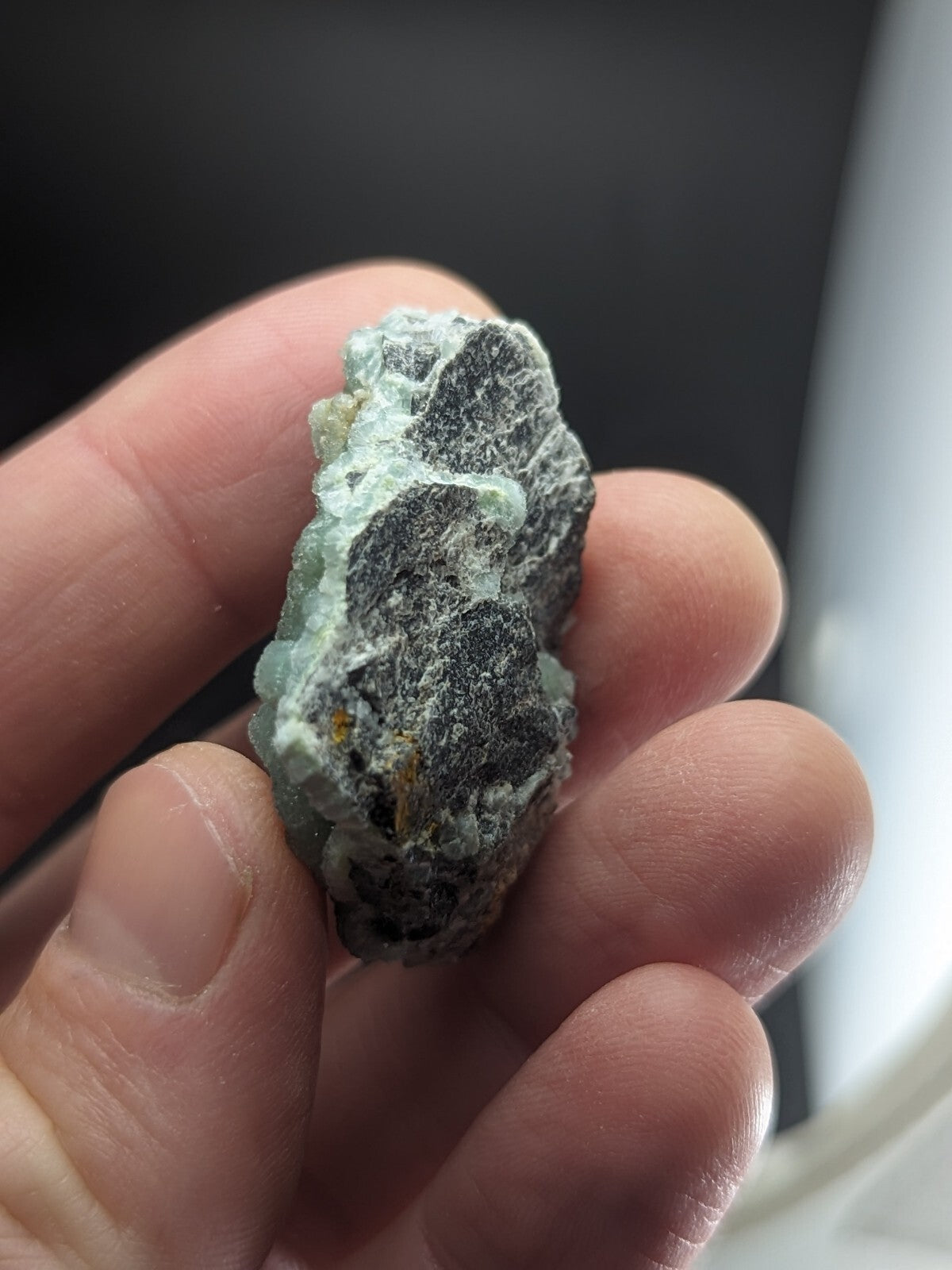 A hand holds a rough, irregularly shaped piece of greenish-blue mineral with dark gray veining and patches of white crystalline material.