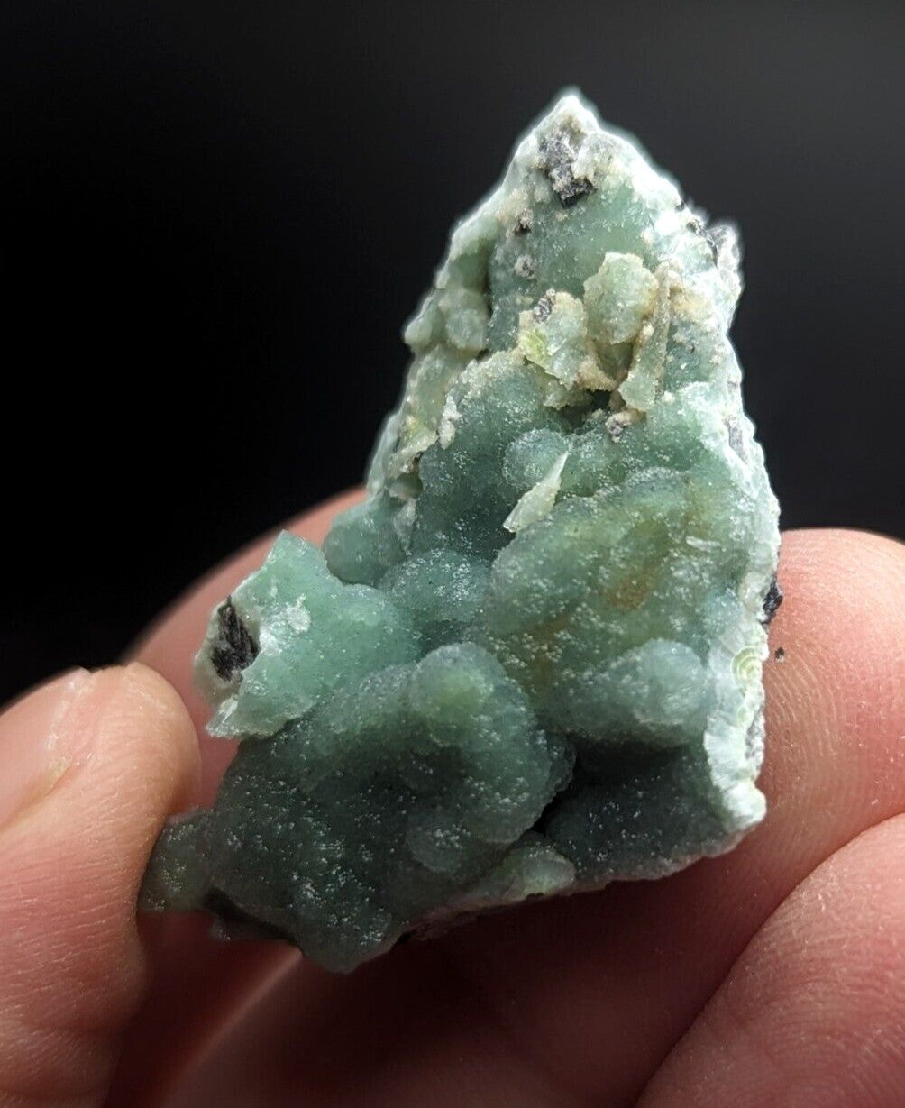 A hand holds a rough, pale green mineral specimen with clustered crystalline formations and patches of white and dark matrix.