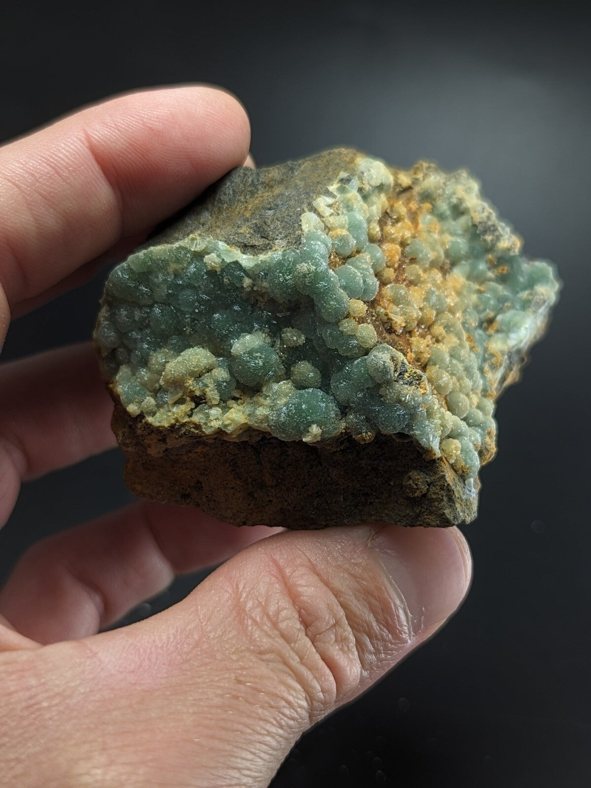 A hand holds a rough, irregularly shaped mineral specimen featuring clusters of translucent green crystals interspersed with patches of yellowish and brown material.