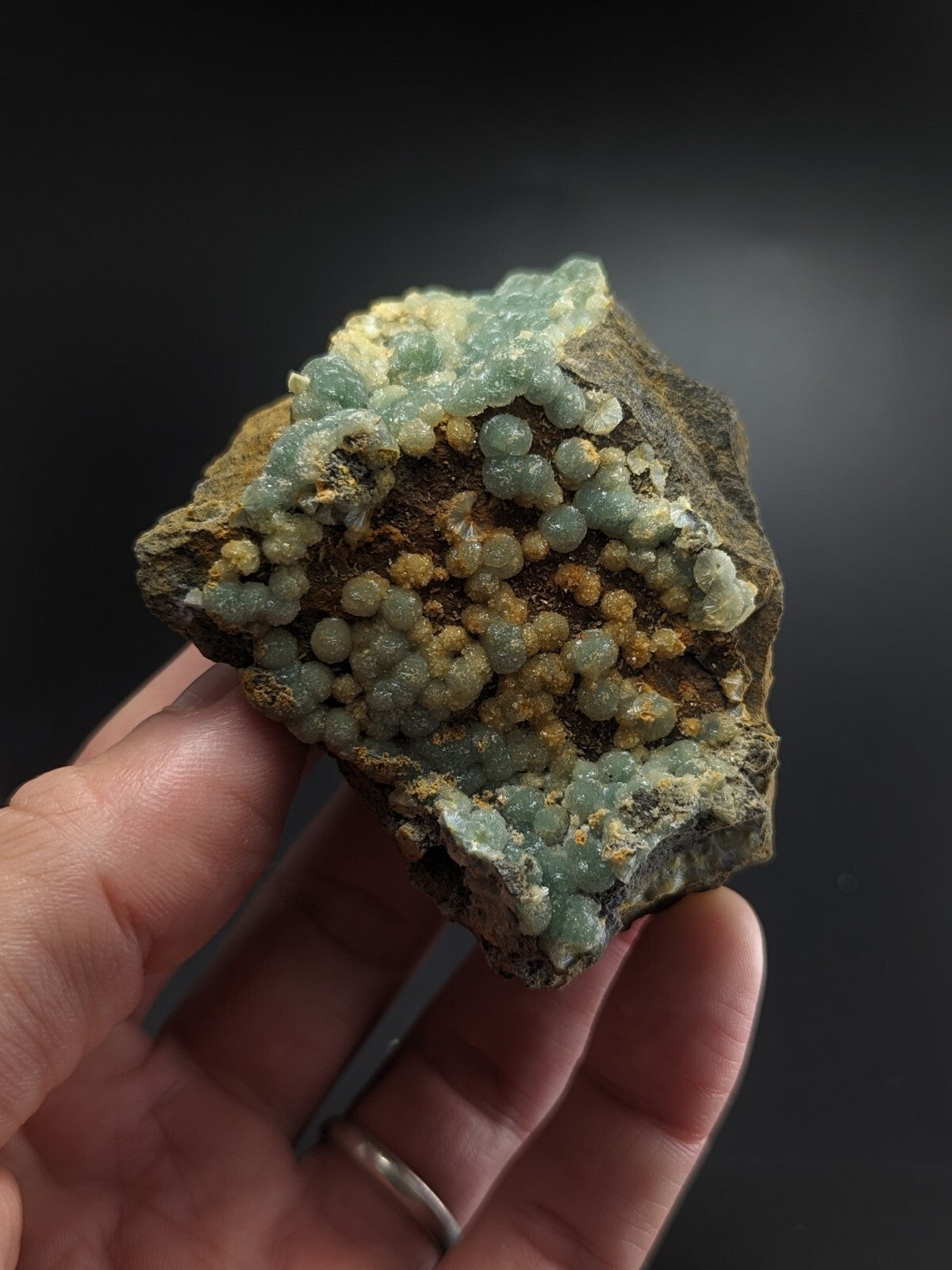 A hand holds a geological specimen featuring clusters of translucent, light green and pale yellow spherical crystals growing from a dark brown rock matrix.