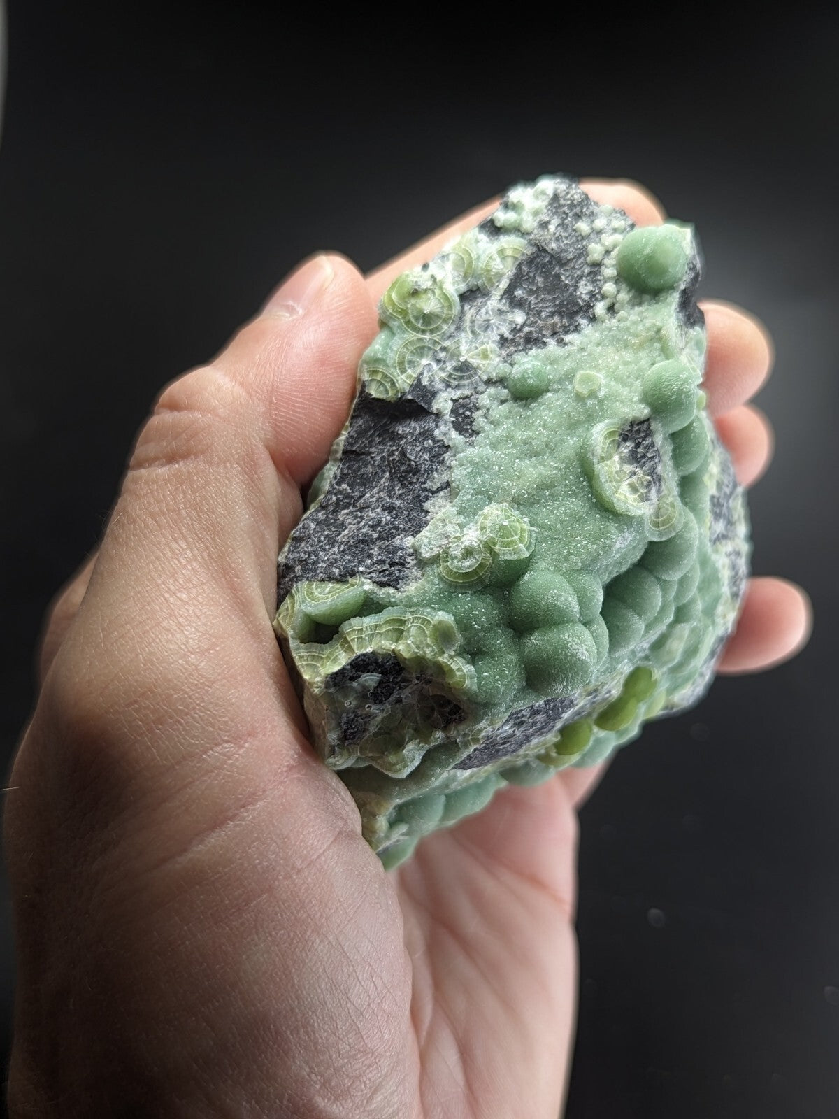 A hand holds a rough, irregularly shaped green and black mineral specimen with bulbous, textured growths.