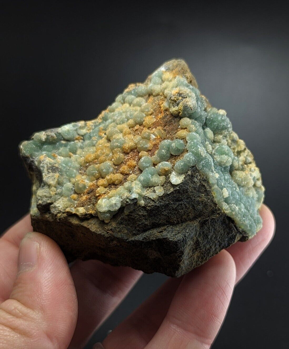 A hand holds a rugged, dark rock specimen featuring clusters of translucent light green and golden-yellow mineral formations.