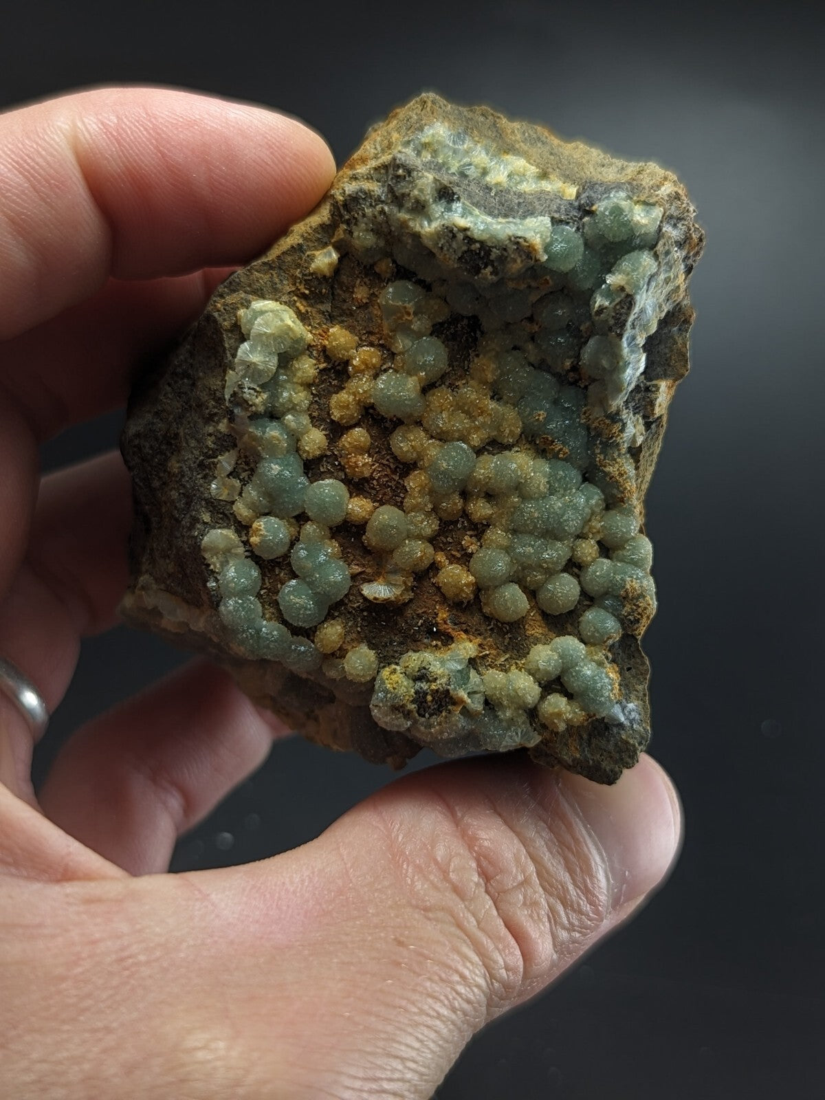 A hand holds a rough, irregularly shaped rock specimen featuring clusters of translucent greenish-blue and yellow spherical crystals growing from its surface.