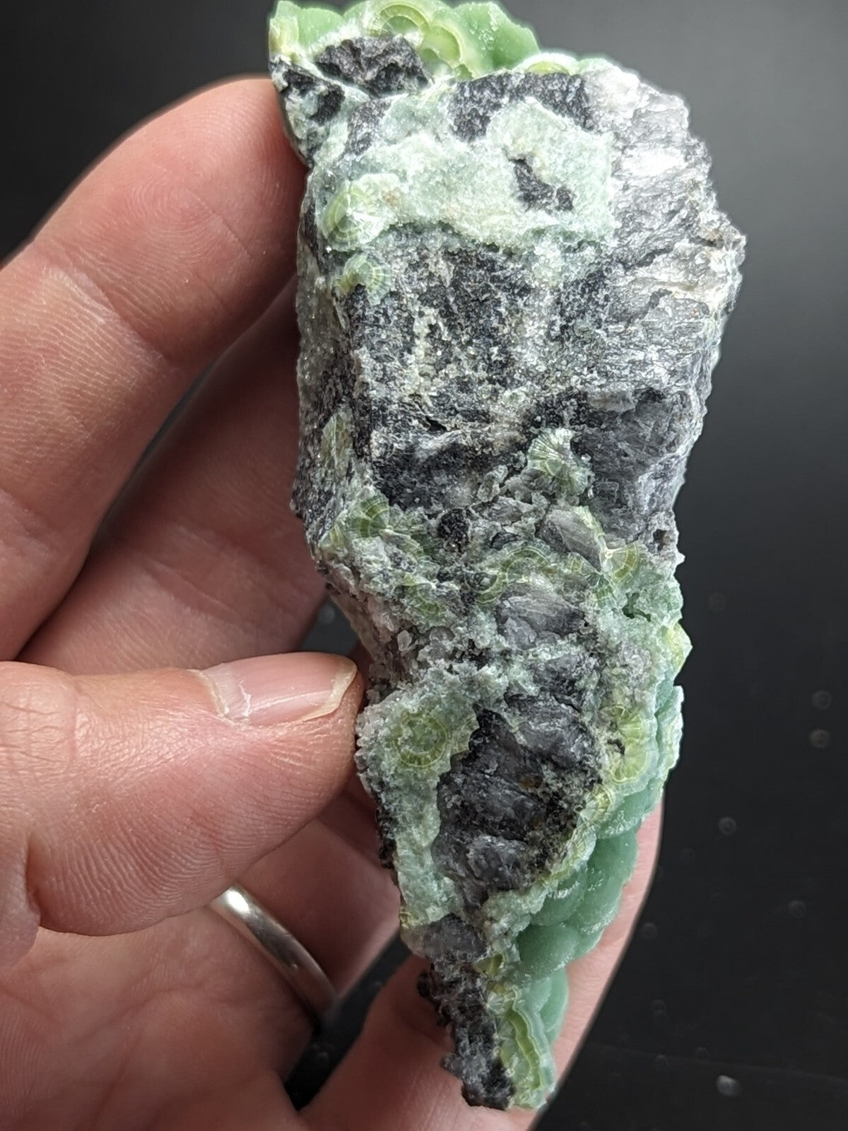 A hand holds a rough, irregularly shaped piece of green and black mineral with visible crystalline textures.