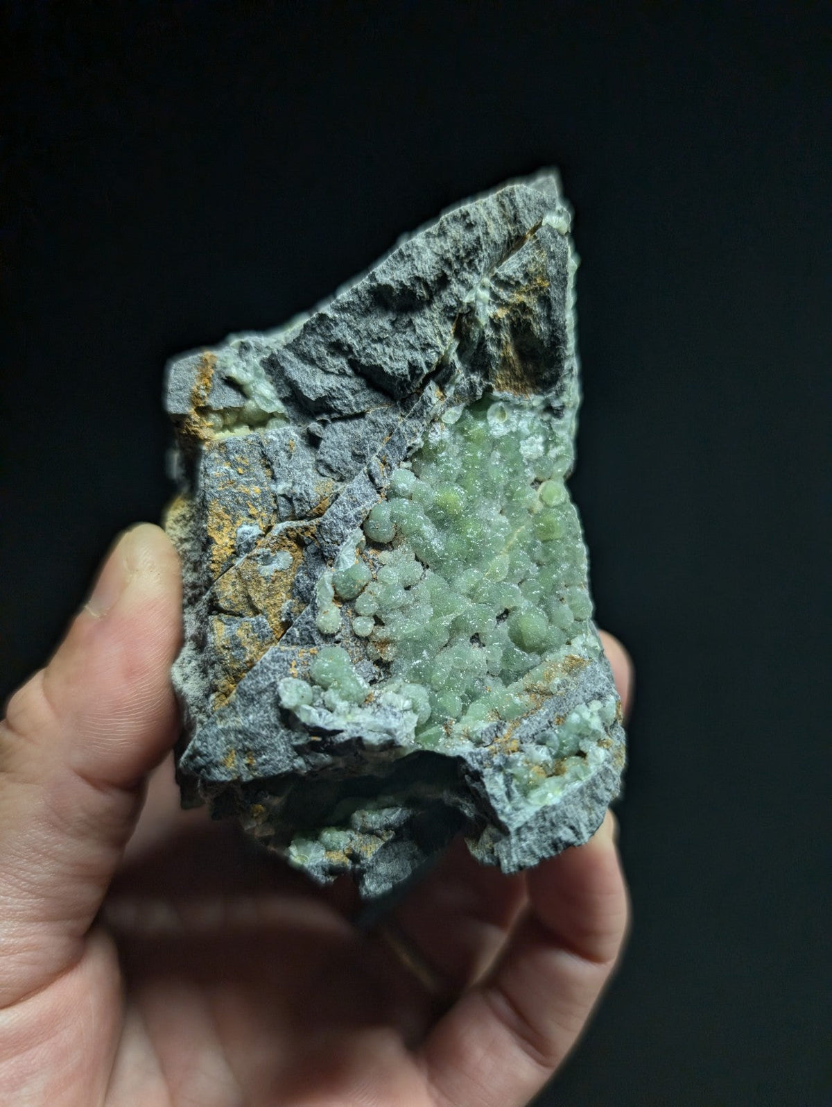 A hand holds a rough, irregularly shaped mineral specimen featuring clusters of translucent green crystals embedded in a grayish matrix with patches of yellow and brown oxidation.
