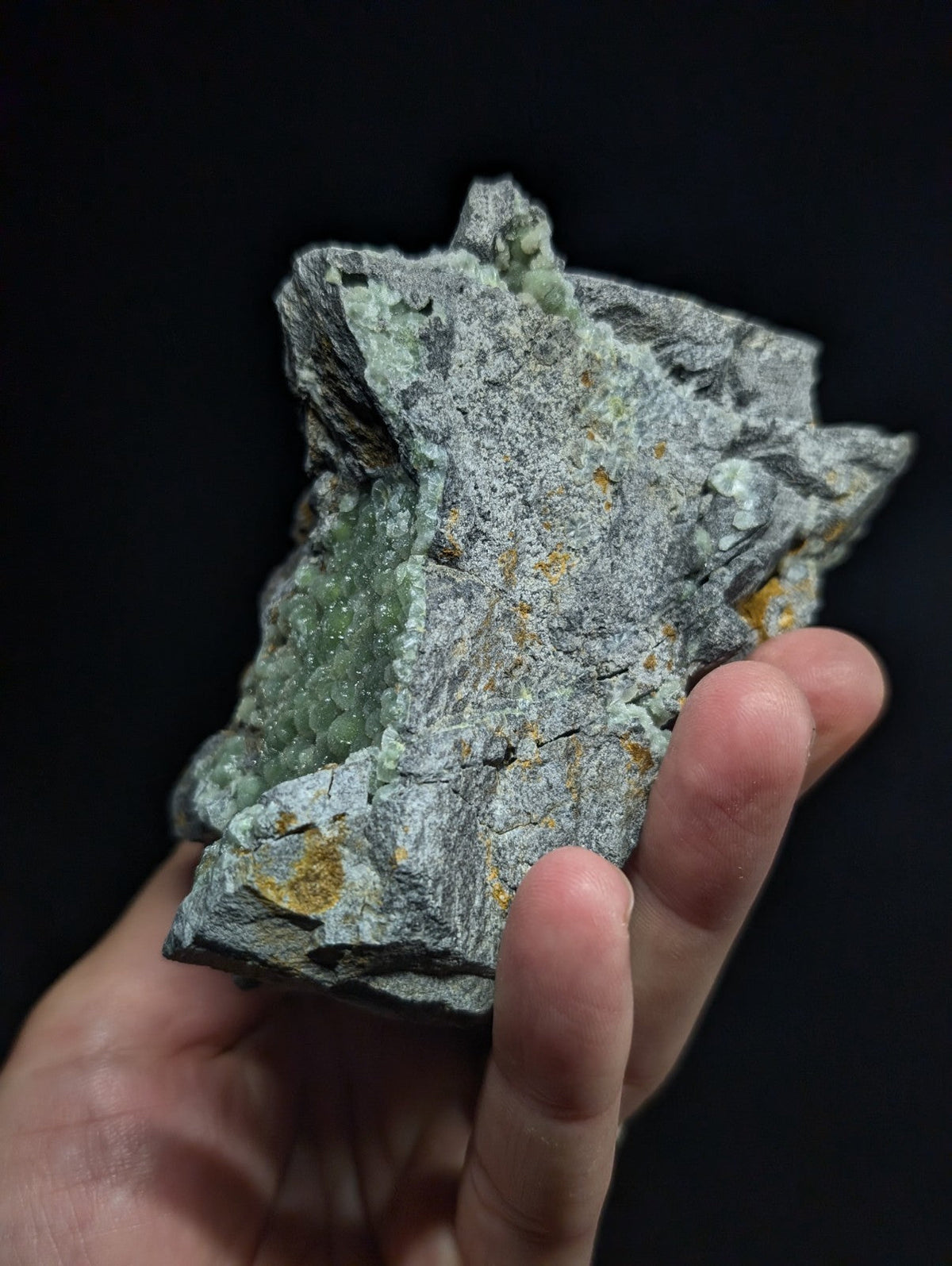 A hand holds a rough, irregularly shaped mineral specimen featuring a gray matrix with patches of translucent green crystals and small yellowish inclusions.