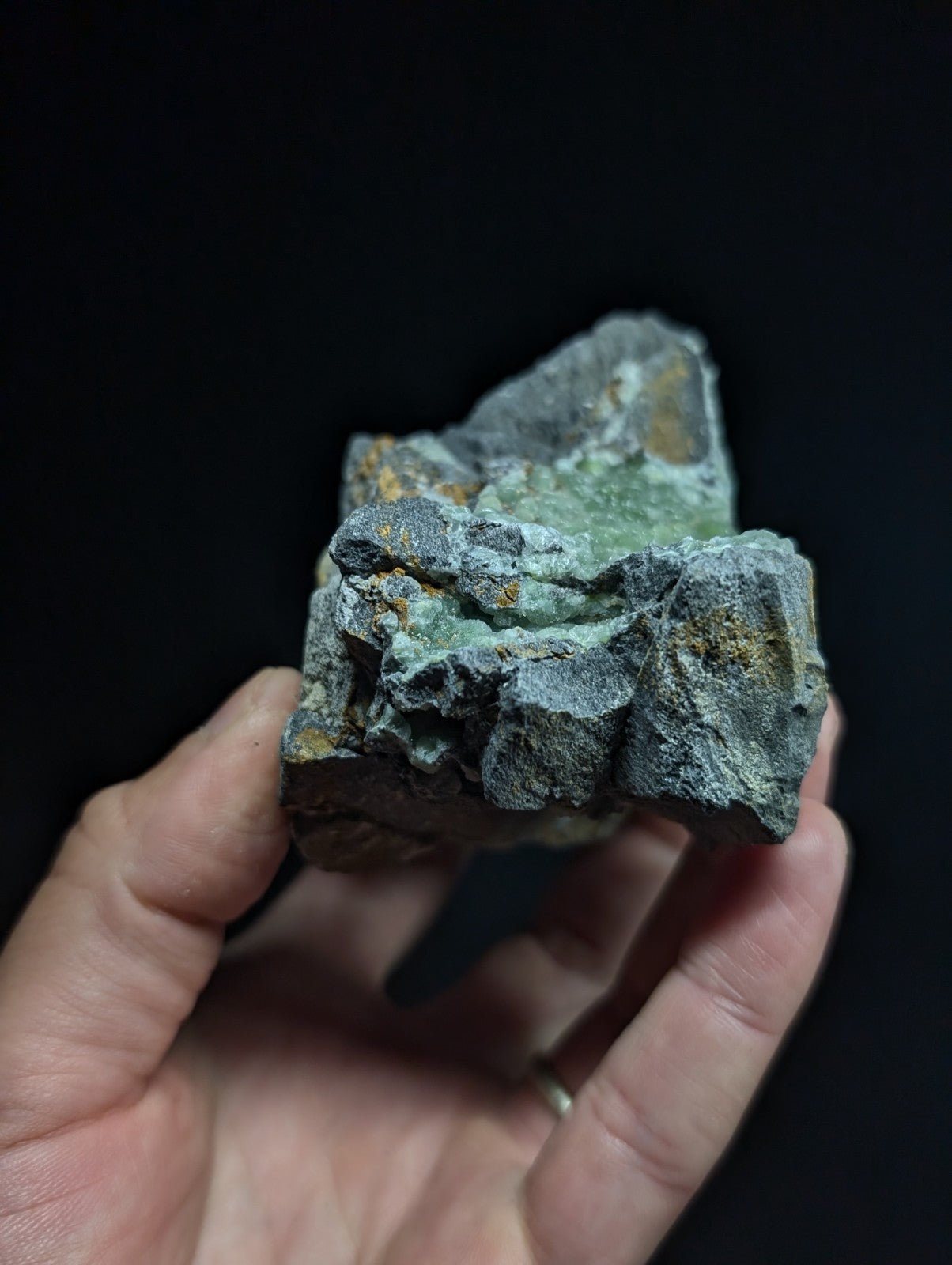 A rough, irregularly shaped mineral specimen with a mottled green and gray surface accented by patches of yellowish-orange patina.