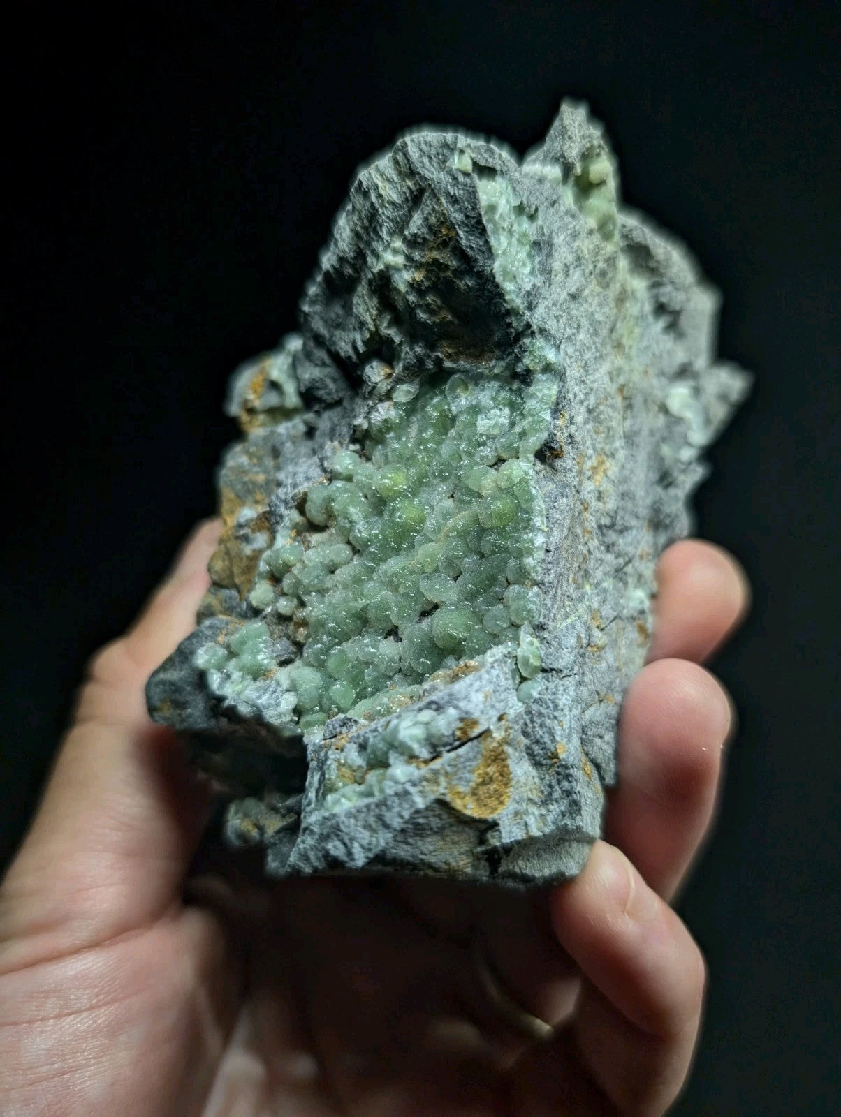 A hand holds a rough, grayish rock specimen featuring clusters of translucent green crystals growing from its surface.