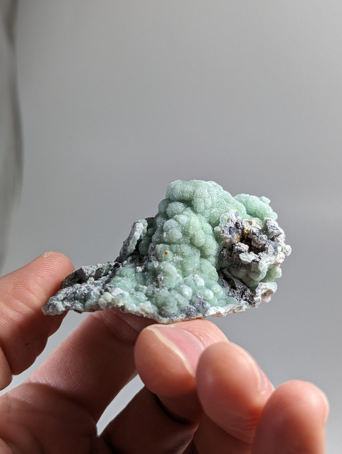 A delicate, light green mineral specimen with a textured, bumpy surface and patches of grayish matrix.