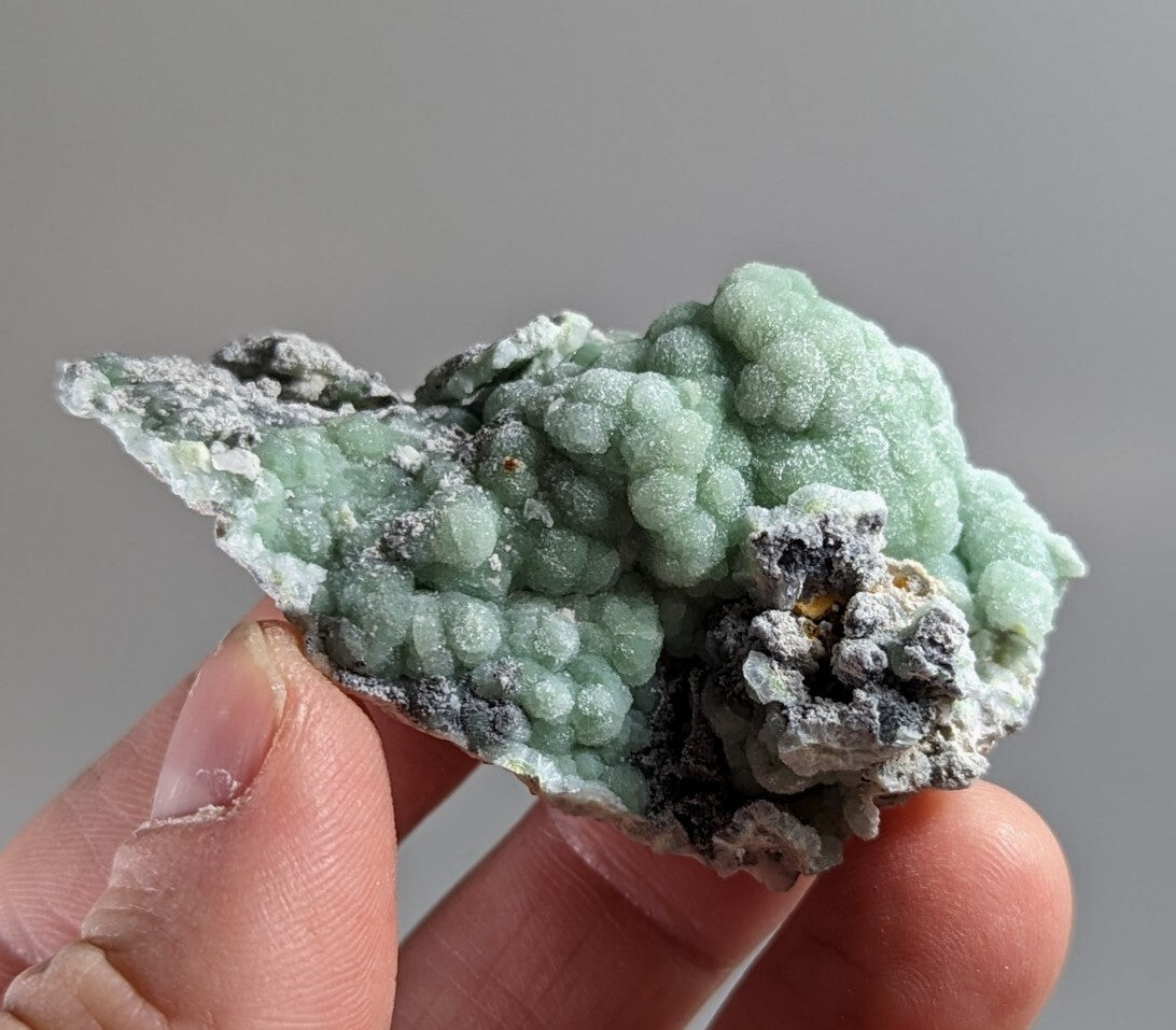 A hand holds a light green, bumpy mineral specimen with grayish-white crystalline patches and some darker inclusions.