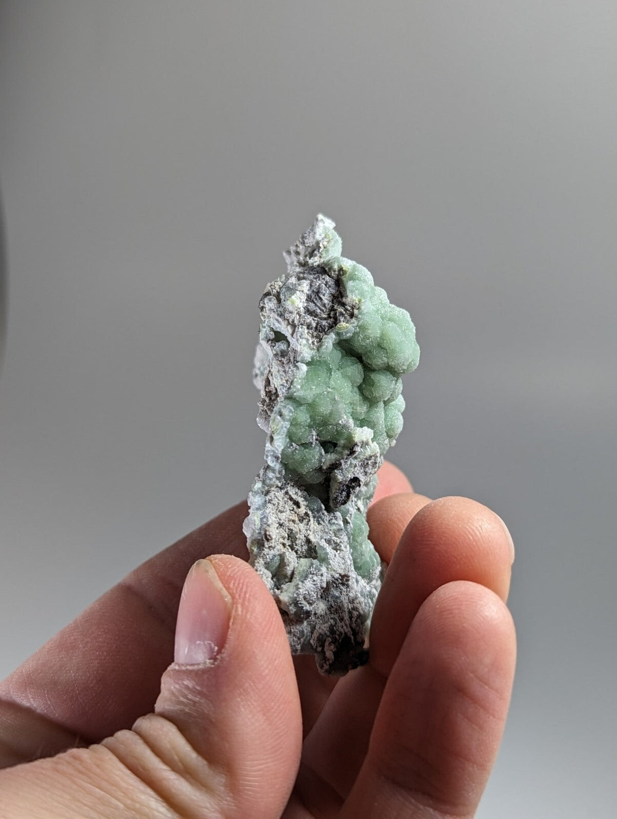 A hand holds a striking green mineral specimen with a textured, bumpy surface and patches of grayish matrix.