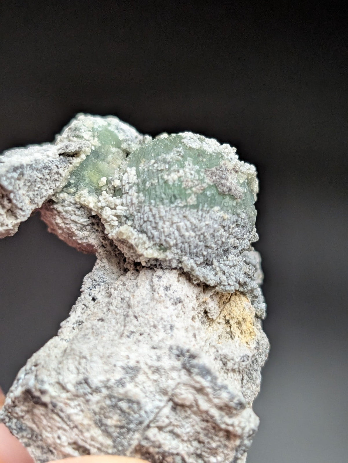 A rough, naturally occurring green fluorite crystal embedded in a gray matrix with some yellowish mineral patches.