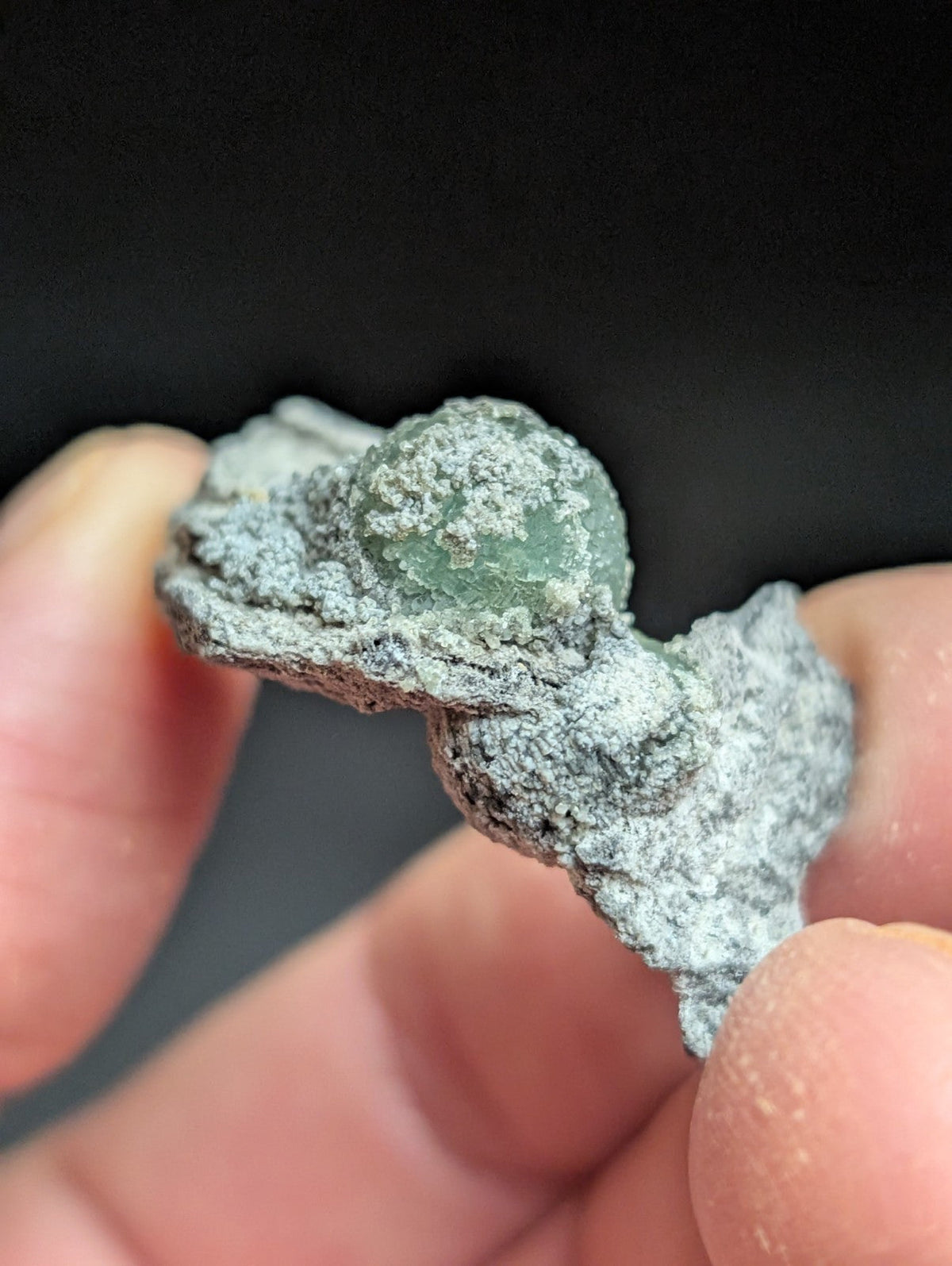 A weathered, gray-green mineral specimen with a rough, textured surface and a translucent green sphere embedded in its center.