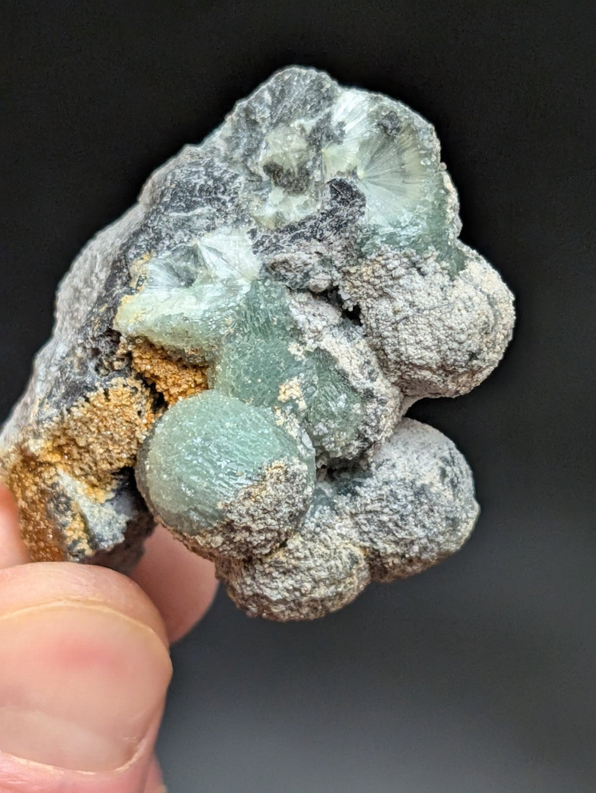 A hand holds a natural mineral specimen featuring translucent green crystals with intricate, rounded formations set against a rough gray matrix and accented by patches of orange-brown material.