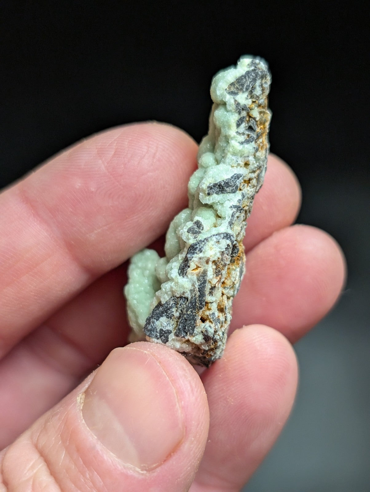 A textured, light green mineral specimen with dark gray and rusty orange patches held in a hand.