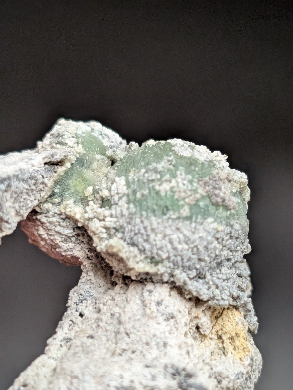 A translucent green mineral specimen embedded in a rough, grayish-white matrix with subtle yellow and reddish-brown patches.