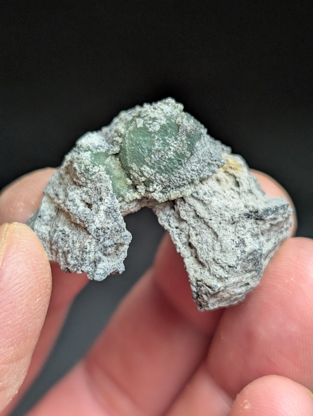 A rough, grayish-green mineral specimen with a visible translucent green gemstone embedded in its rocky matrix.
