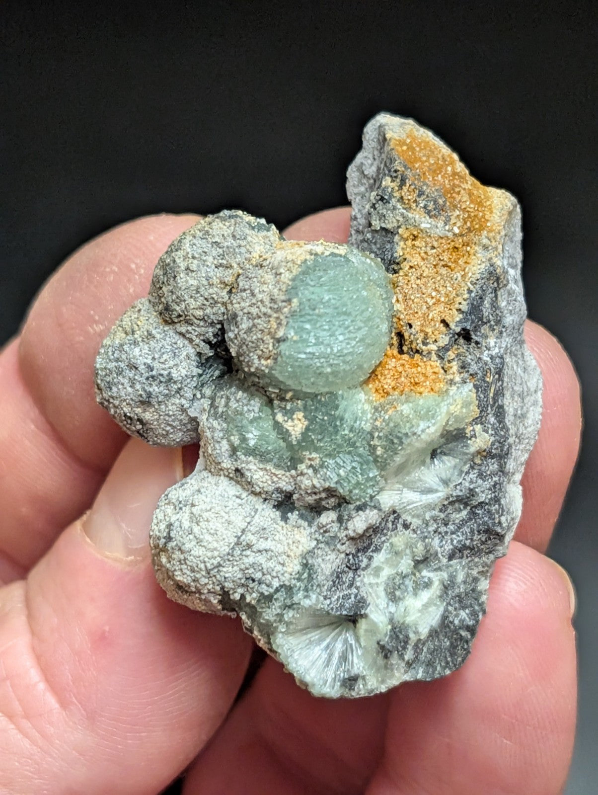 A hand holds a natural mineral specimen featuring translucent green crystals nestled within gray, textured rock and accented with patches of orange-yellow material.
