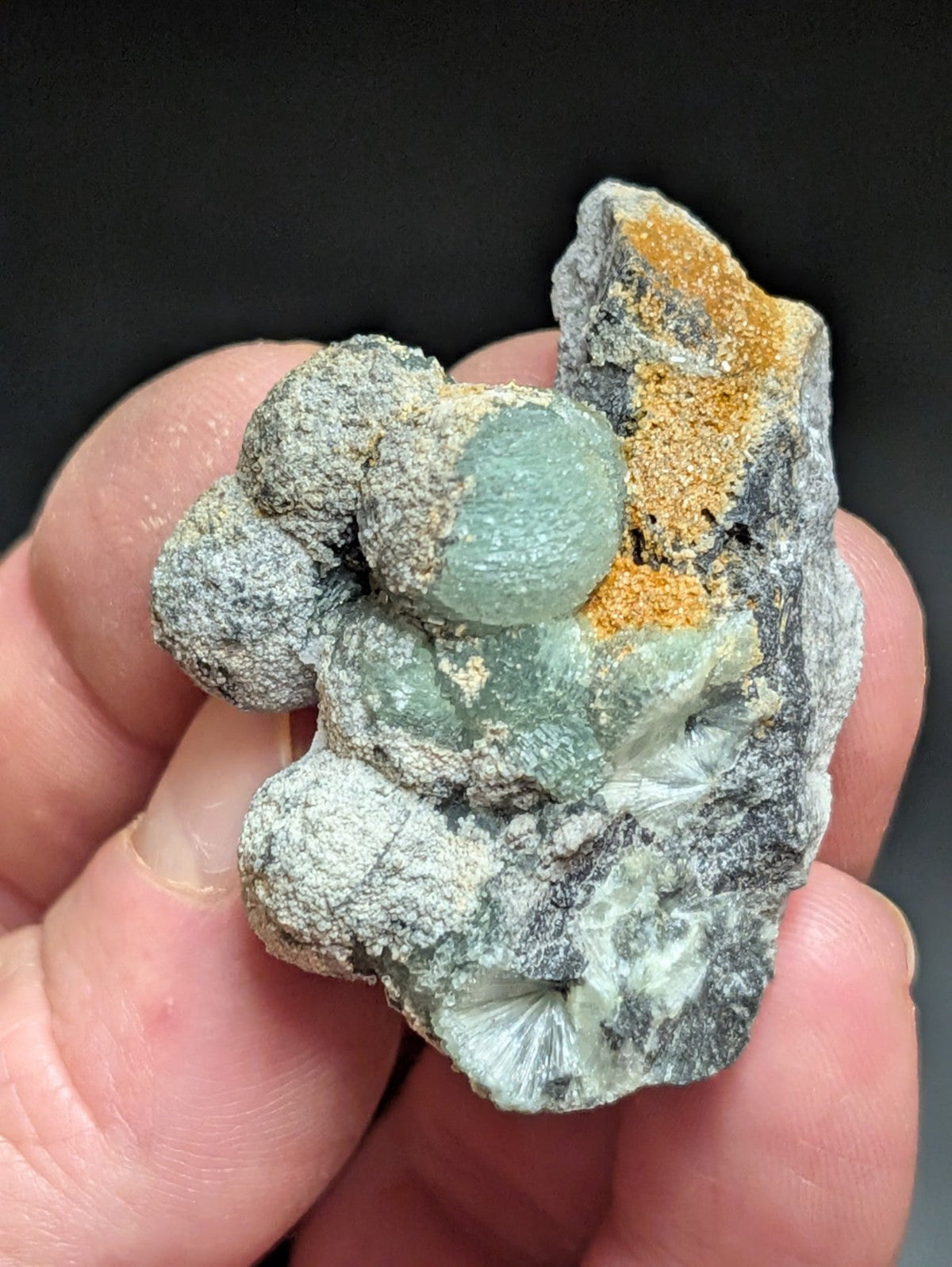 A hand holds a natural mineral specimen featuring translucent green crystals nestled within gray, textured rock and accented with patches of orange-yellow material.