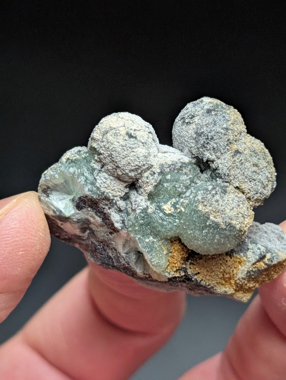 A hand holds a cluster of translucent greenish-blue crystals with rounded, bulbous formations coated in a fine gray powdery substance.