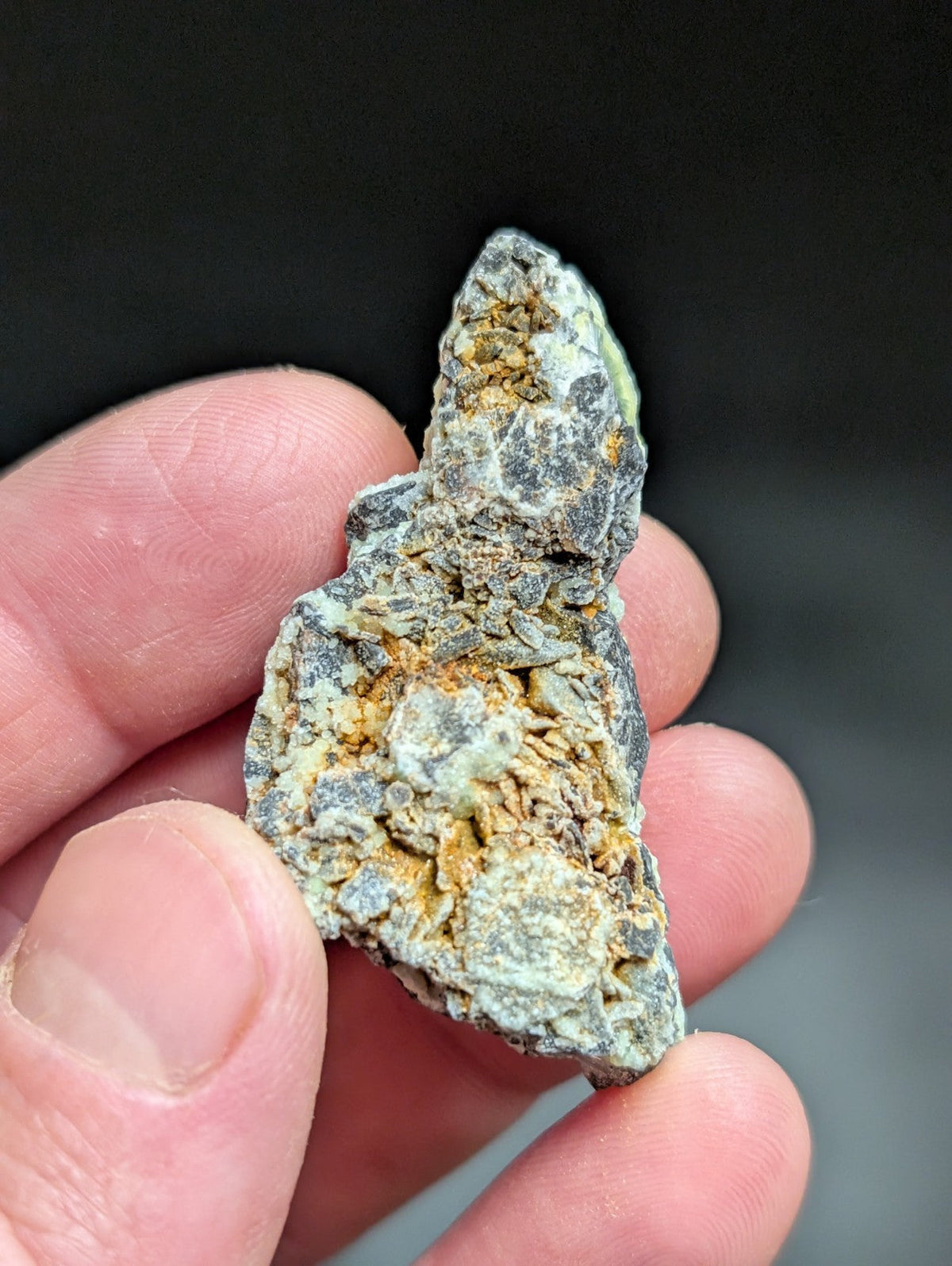 A rough, irregularly shaped mineral specimen with a mottled surface of gray, greenish-blue, and yellow-orange hues.