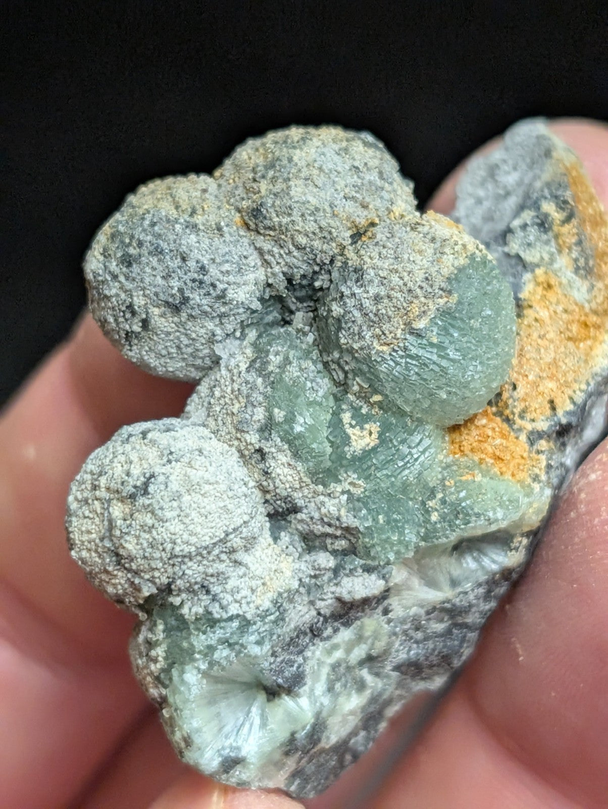 A hand holds a natural mineral specimen featuring rounded, translucent green crystals embedded in a matrix of gray and orange-yellow earthy material.