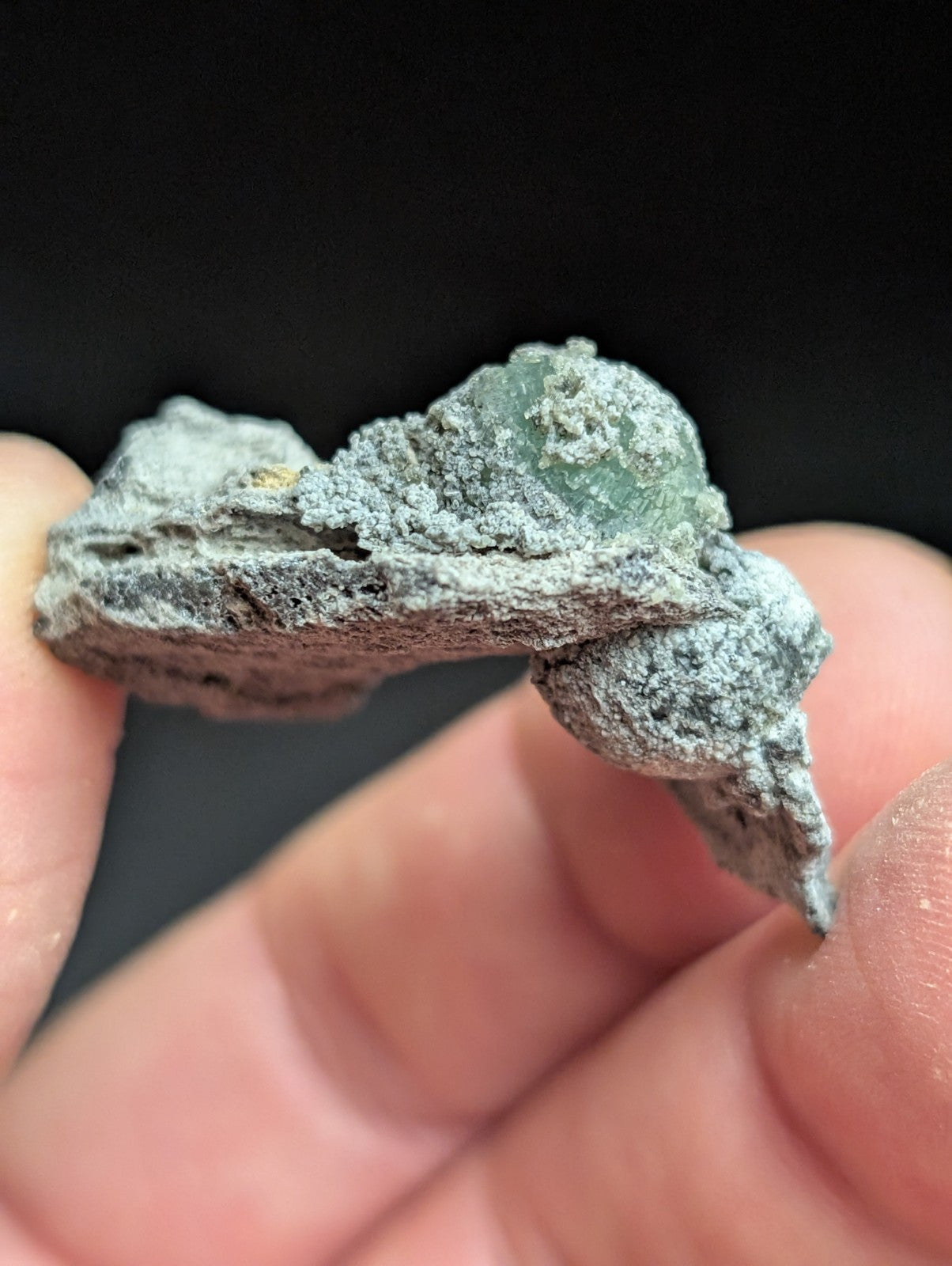 A weathered, grayish-green mineral specimen with a rough, textured surface and visible crystalline formations.