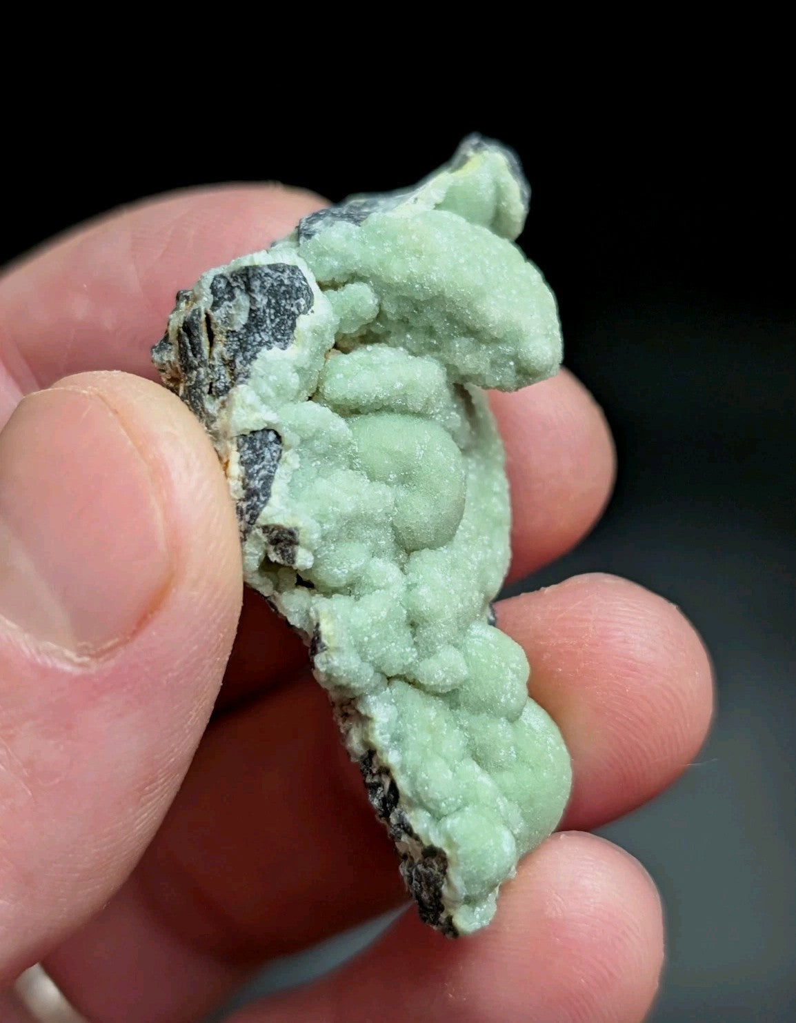 A hand holds a light green, textured mineral specimen with dark gray rock matrix.