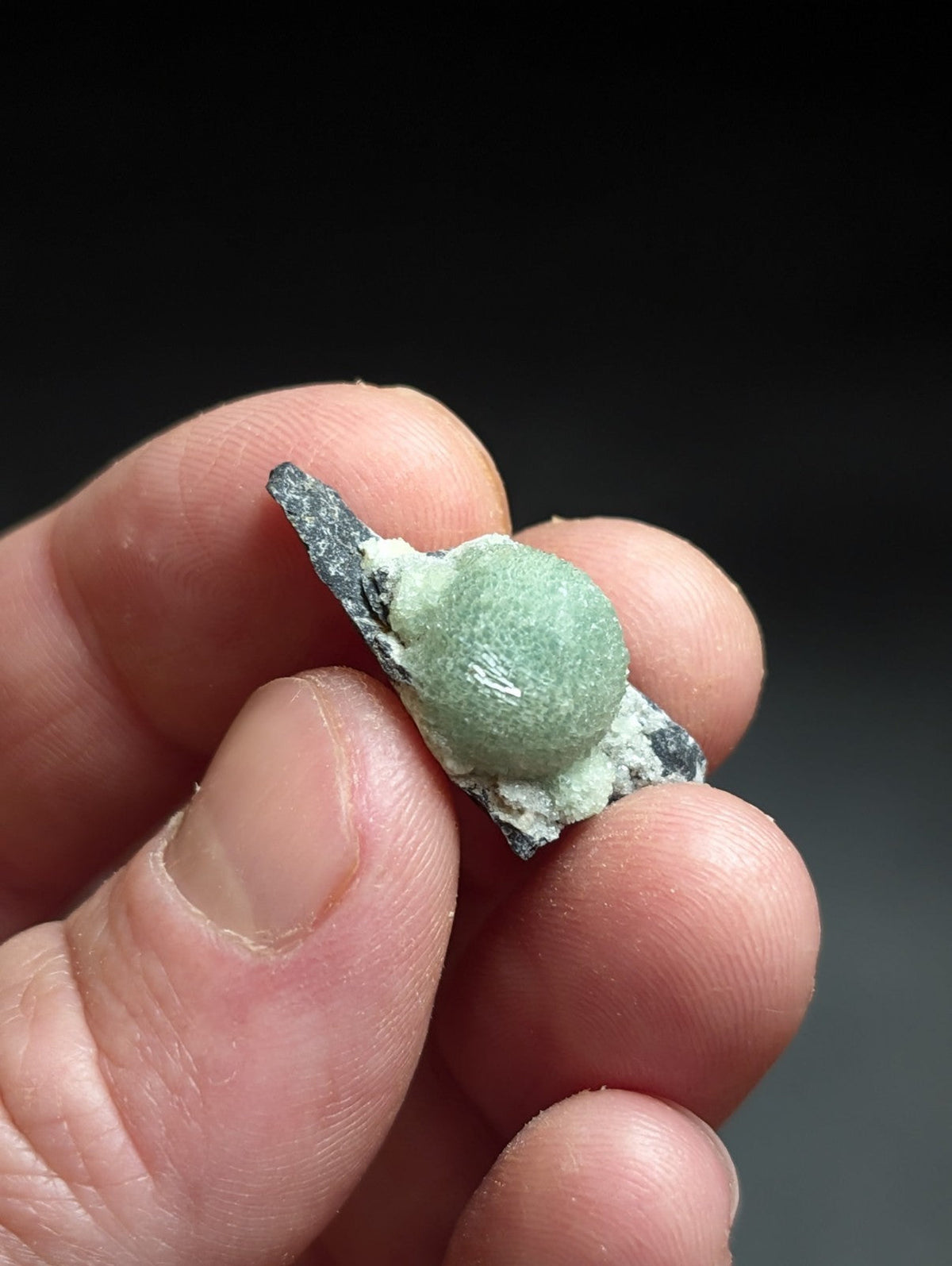 A small, round, light green mineral specimen with a slightly rough texture and white crystalline deposits on its surface.