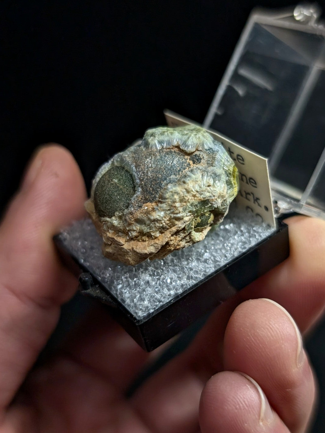 A greenish, rough-textured mineral specimen with a prominent dark circular inclusion nestled in its rocky matrix.