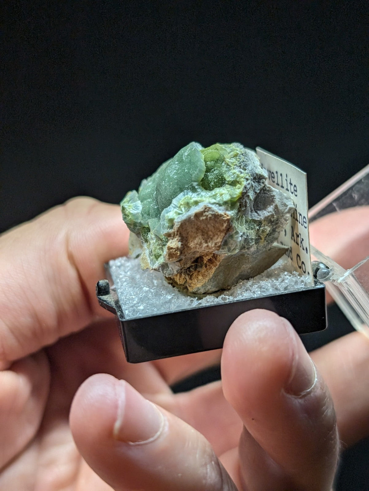 A green, translucent mineral specimen with a crystalline texture and earthy brown matrix, displayed in a black display case.