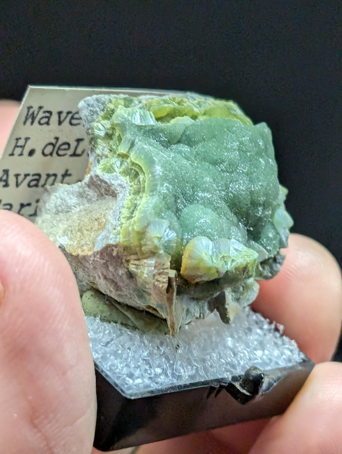 A green and yellow mineral specimen with a rough, textured surface mounted on a clear acrylic display base.