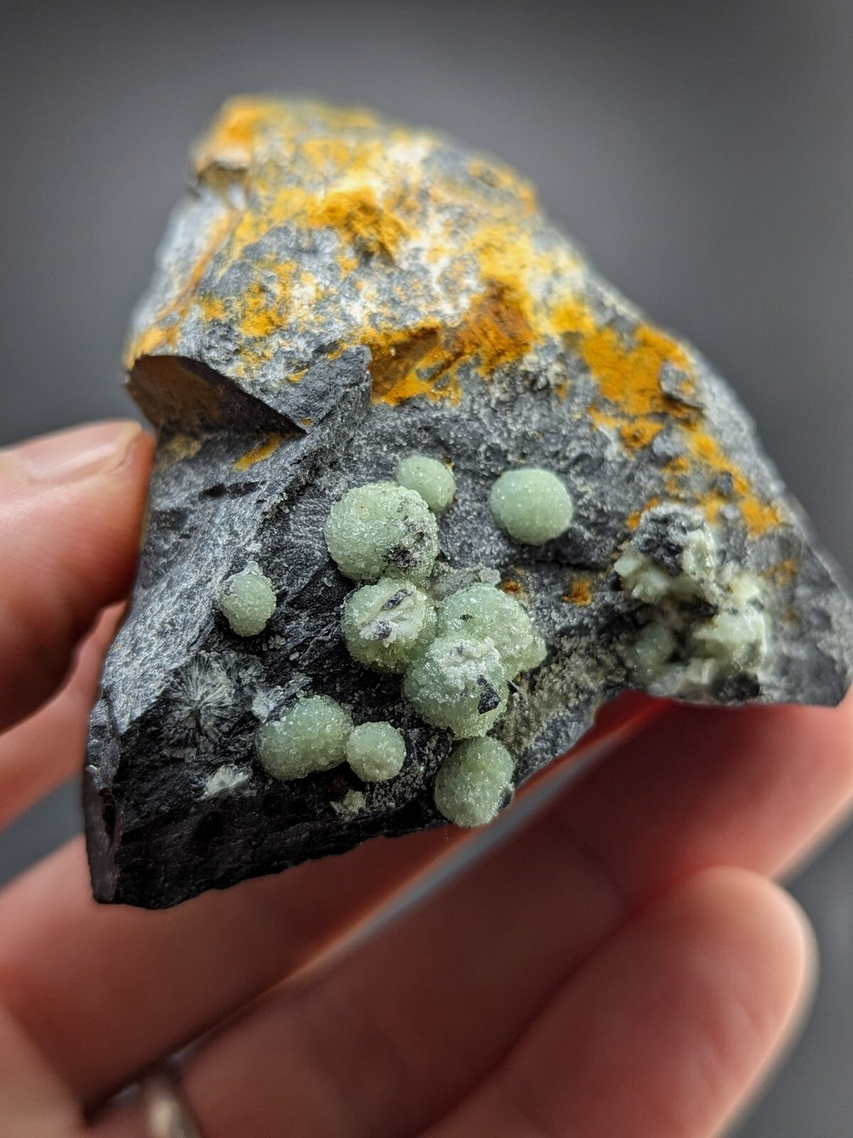 A hand holds a dark gray rock specimen featuring clusters of translucent, light green spherical crystals with patches of vibrant yellow mineral coating.