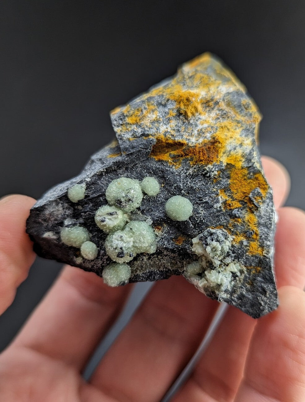 A hand holds a dark gray rock specimen featuring vibrant orange-yellow mineral patches and clusters of translucent, pale green spherical crystals.