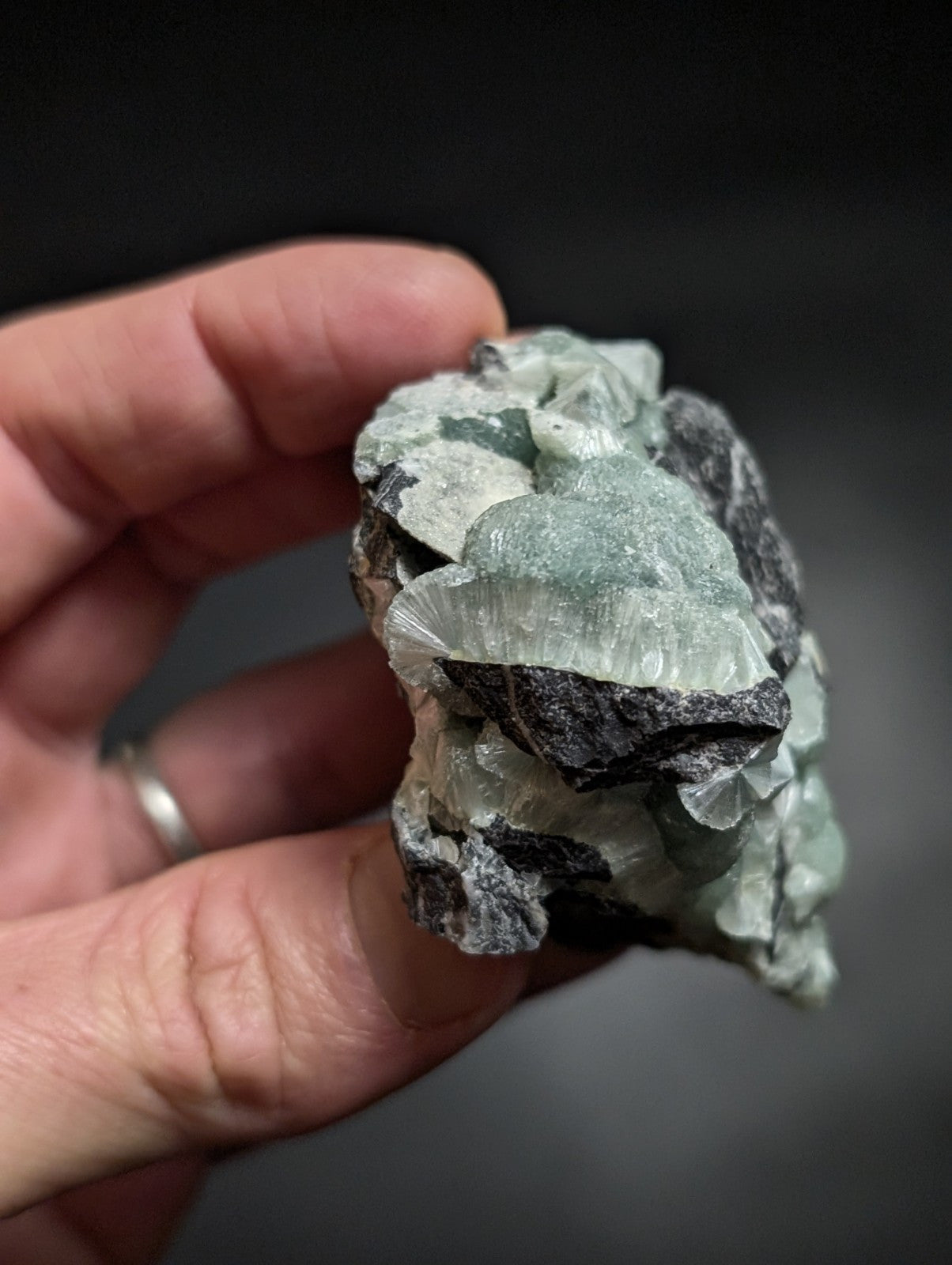 A hand holds a rough, irregularly shaped mineral specimen featuring translucent light green crystals with sharp facets growing from dark gray rock.