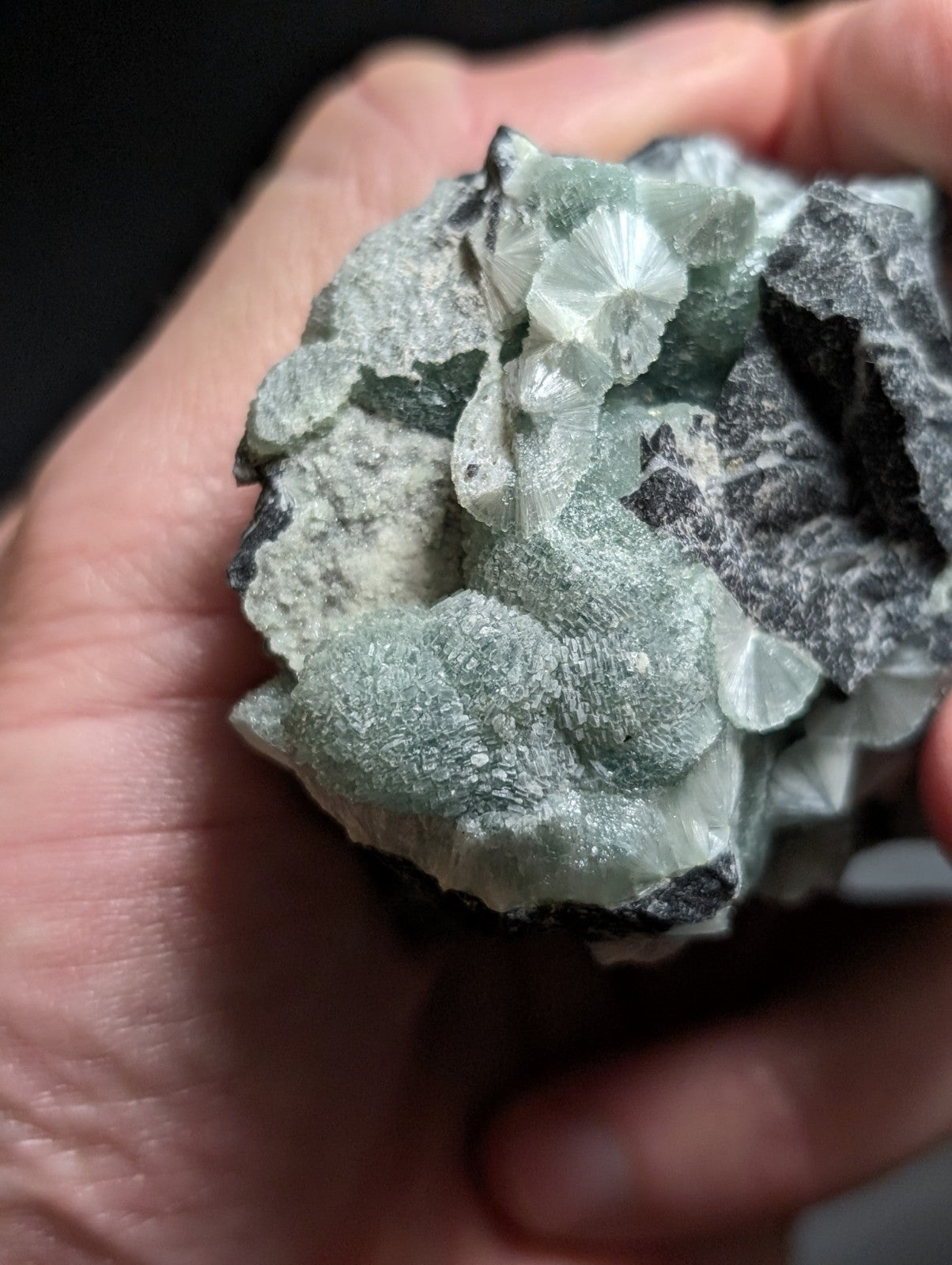 A hand holds a rough, irregularly shaped mineral specimen featuring translucent green crystals with radial facets emerging from a darker gray matrix.