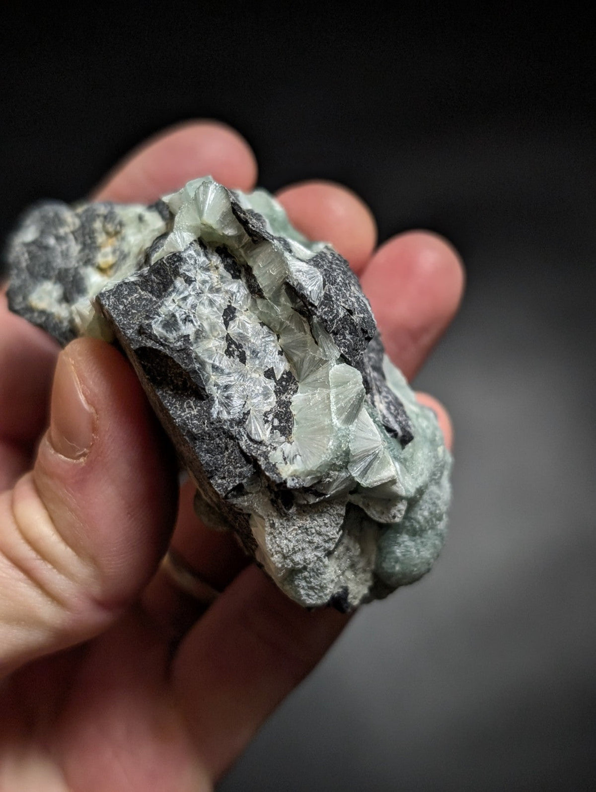 A hand holds a rough, irregularly shaped mineral specimen featuring translucent light green crystals embedded in dark gray rock.