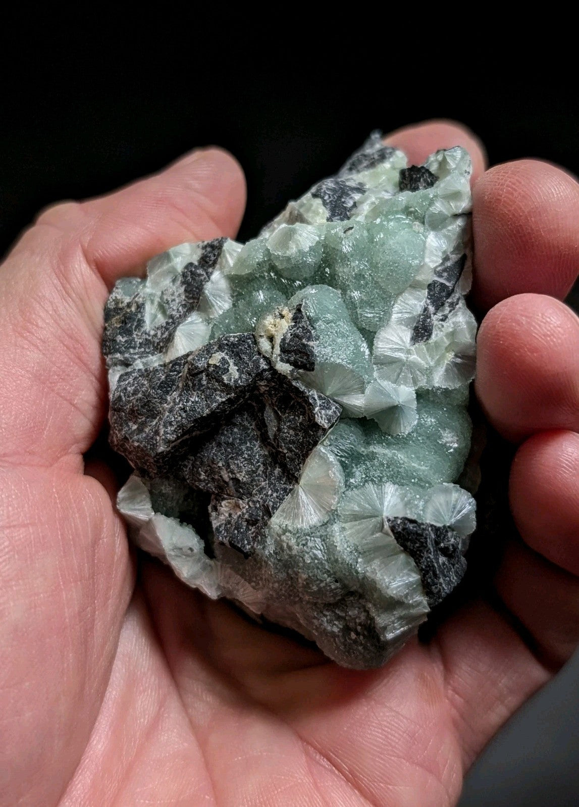 A hand holds a natural green and black mineral specimen with crystalline formations.