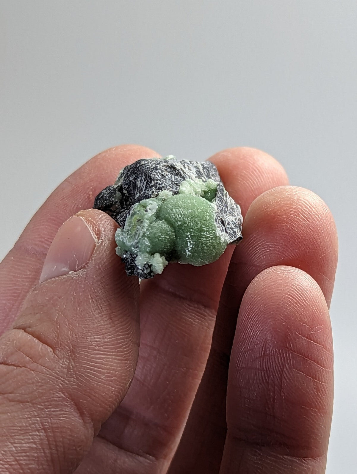A hand holds a small, irregularly shaped mineral specimen featuring vibrant green crystalline formations nestled within dark gray rock.