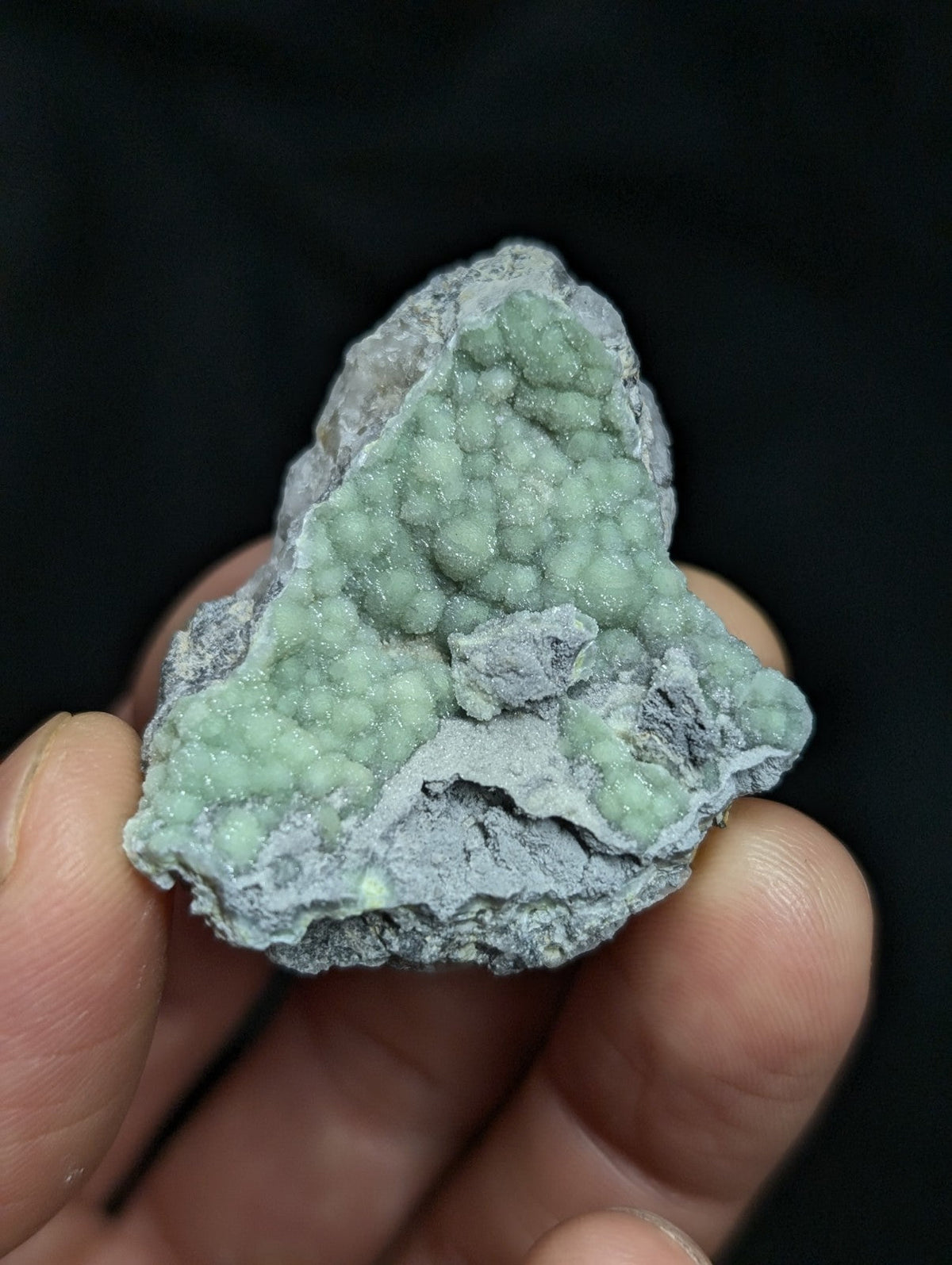 A hand holds a rough, irregularly shaped mineral specimen featuring a striking light green crystalline formation with a textured surface against a dark gray matrix.