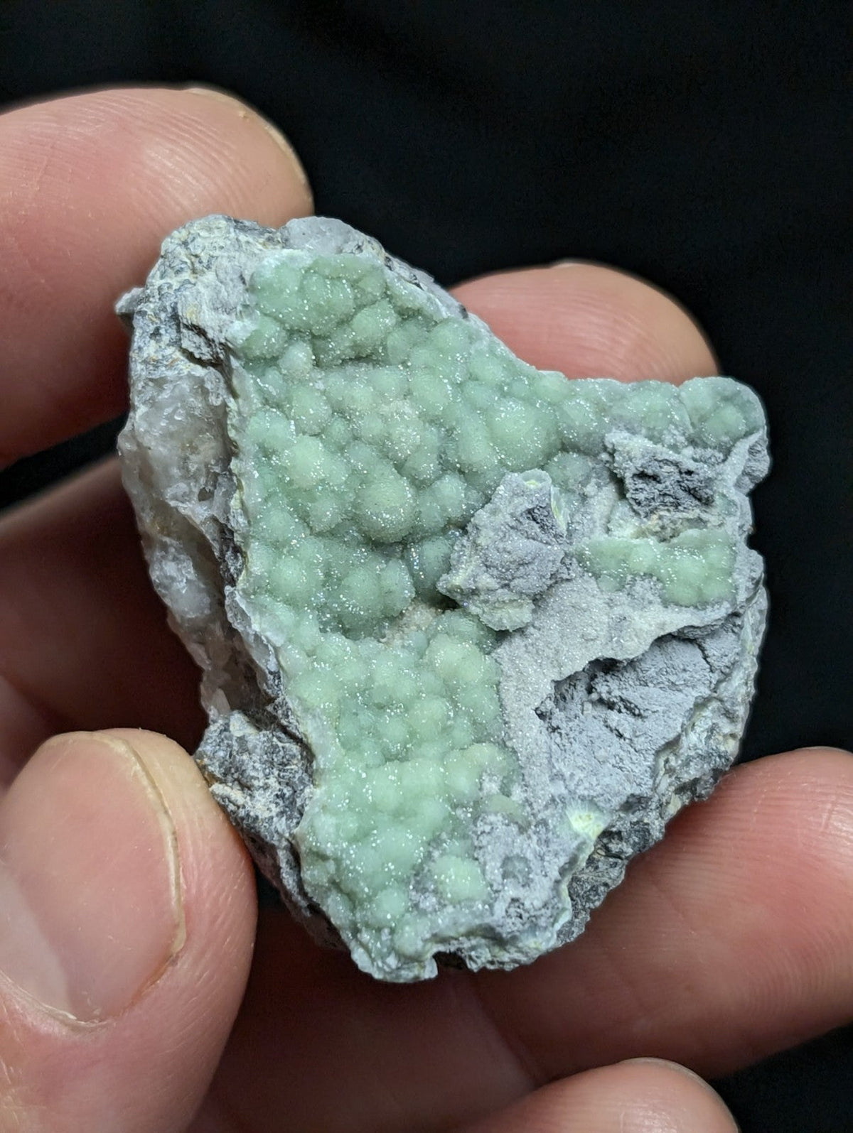 A hand holds a rough, irregularly shaped mineral specimen featuring clusters of pale green, bead-like crystals growing from a grayish matrix.
