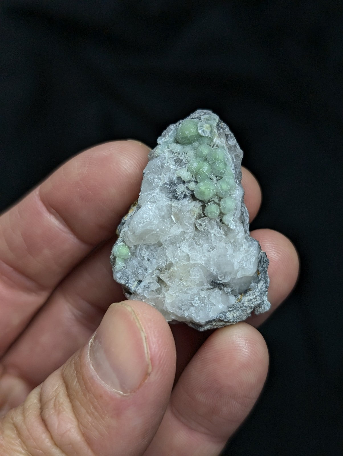 A hand holds a rough, irregularly shaped mineral specimen featuring translucent white crystals with clusters of pale green spherical formations.